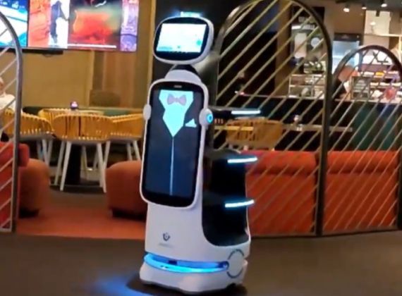 Robot server in a restaurant, wearing a tuxedo on its screen, blue lights, carrying trays.