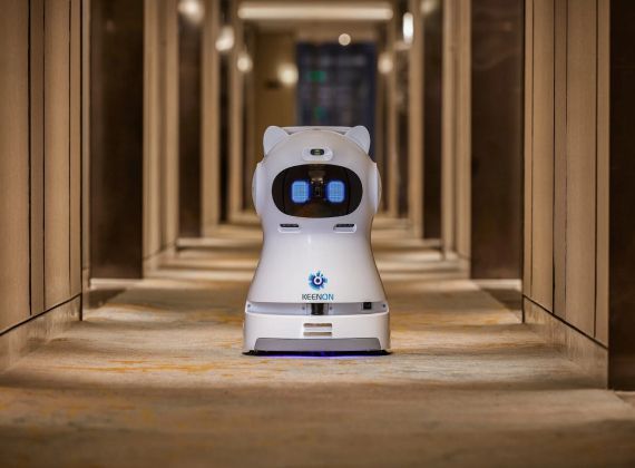 Robot in a hotel hallway; white with blue eye-like screens; delivering items.