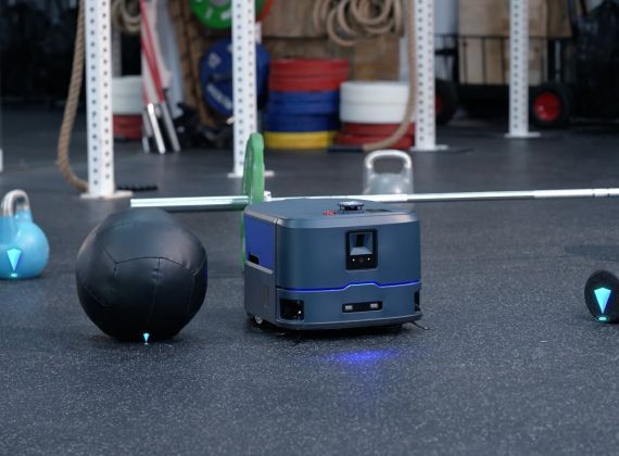 A small, dark gray robot navigating a gym, with workout equipment and vertical poles in the background.