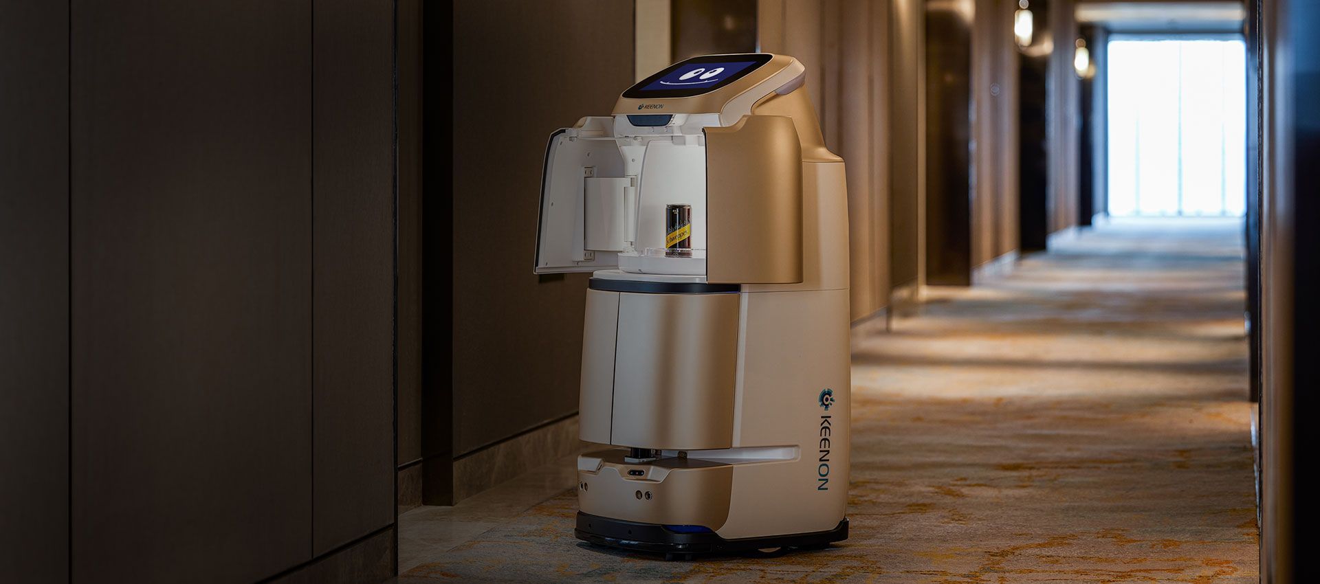 A delivery robot in a hotel hallway. The robot is beige with a screen on top. A doorway is at the end of the hallway.