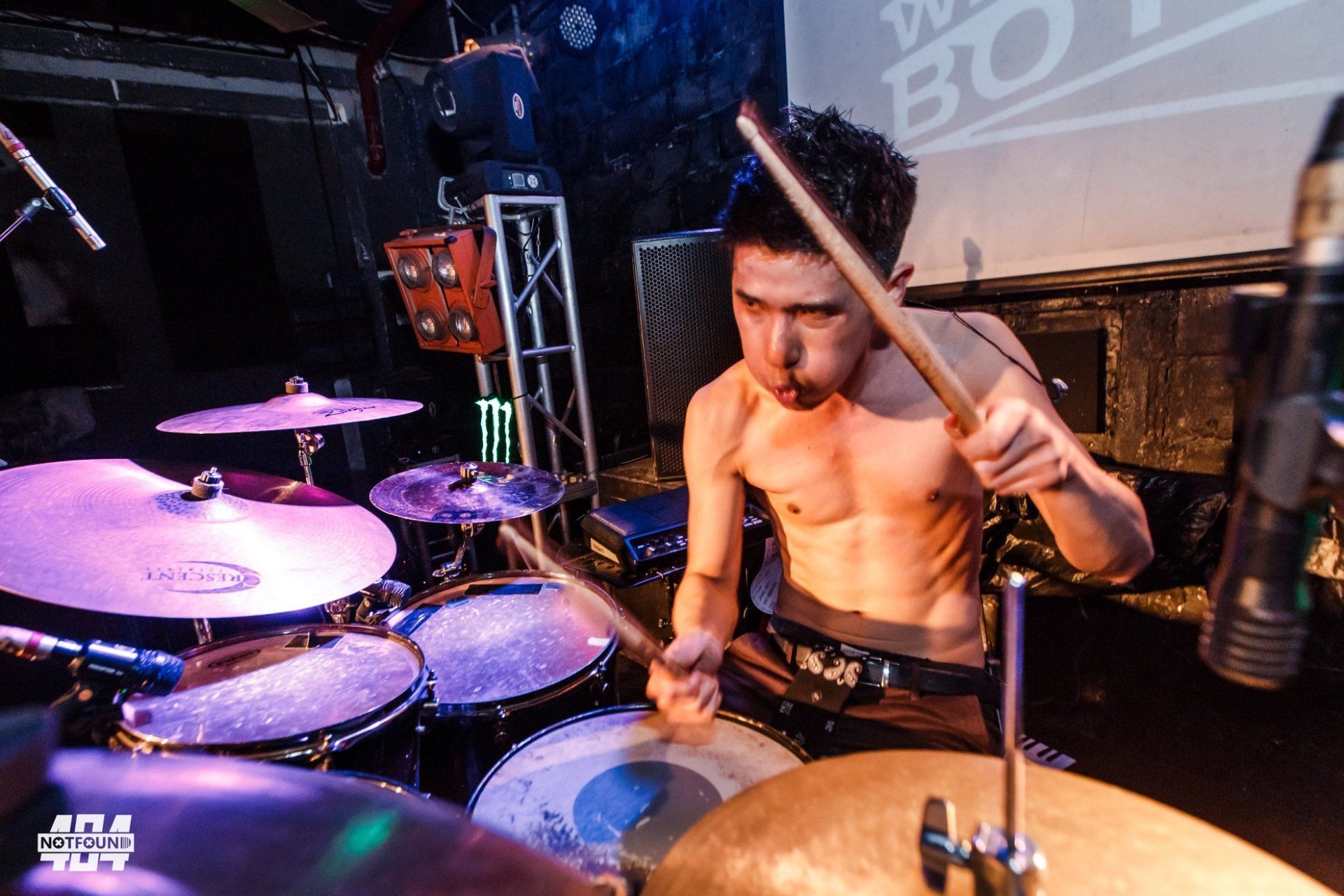 A shirtless man is playing drums in a dark room.
