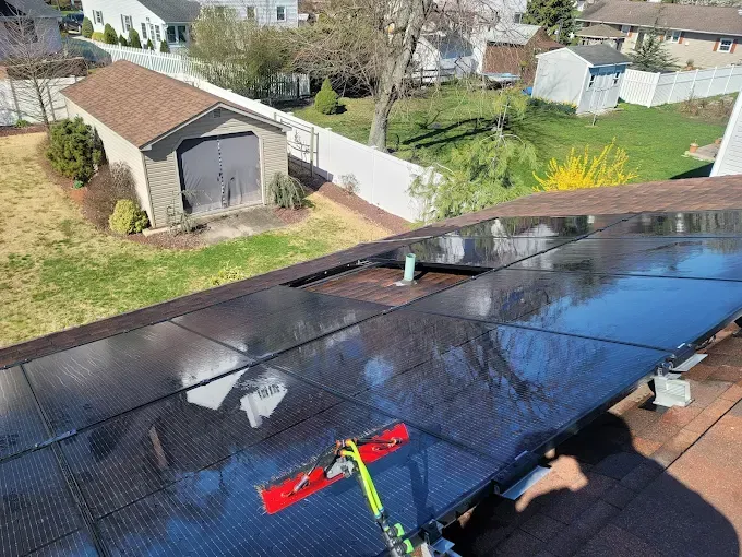 Person cleaning solar panels on a brown shingled roof with a long-handled brush; a white fence and a shed are visible.