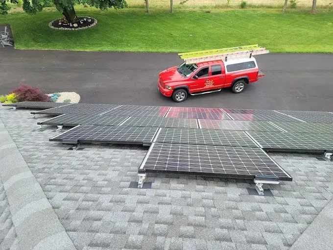 Solar panels installed on a rooftop next to a red pickup truck carrying a ladder on a driveway, green lawn in background.