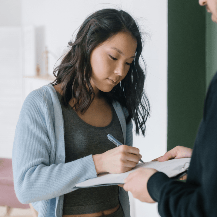 Woman signing a document held by another person in a light-filled room.