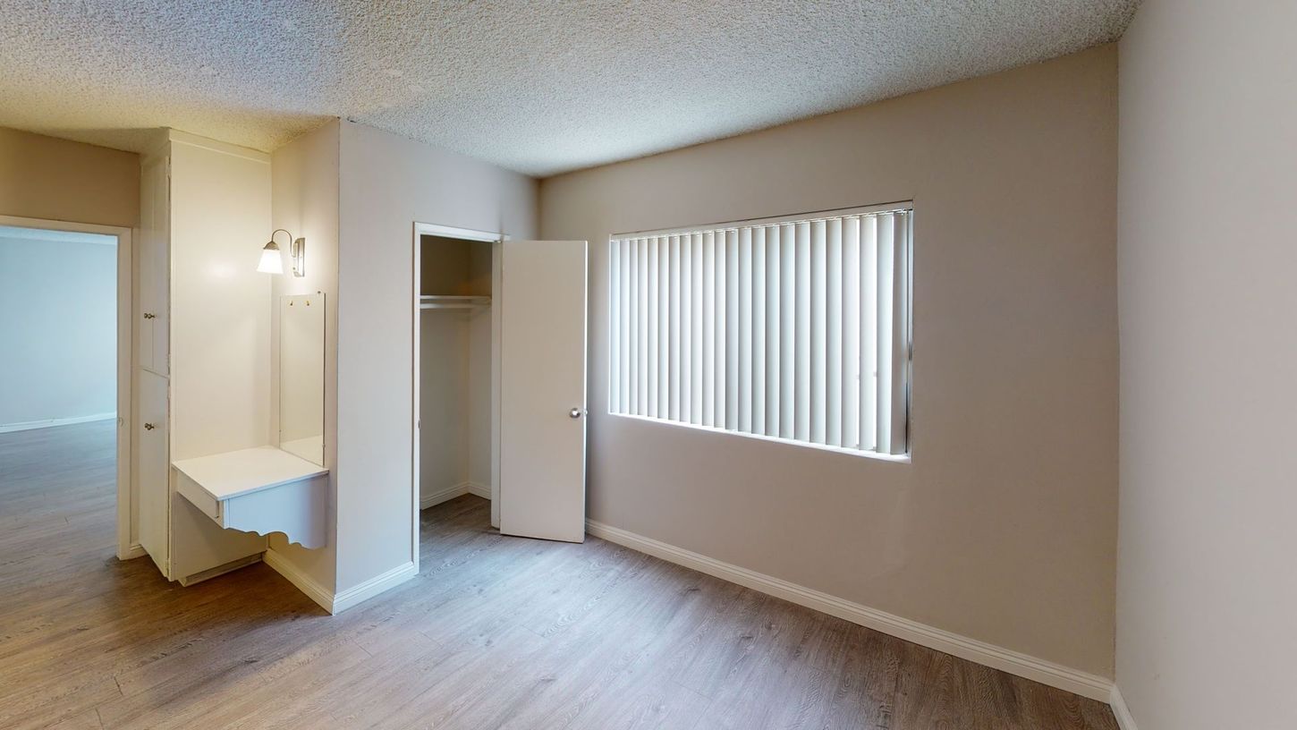 Empty bedroom with closet, window, and small vanity with lights. Beige walls, light wood flooring.