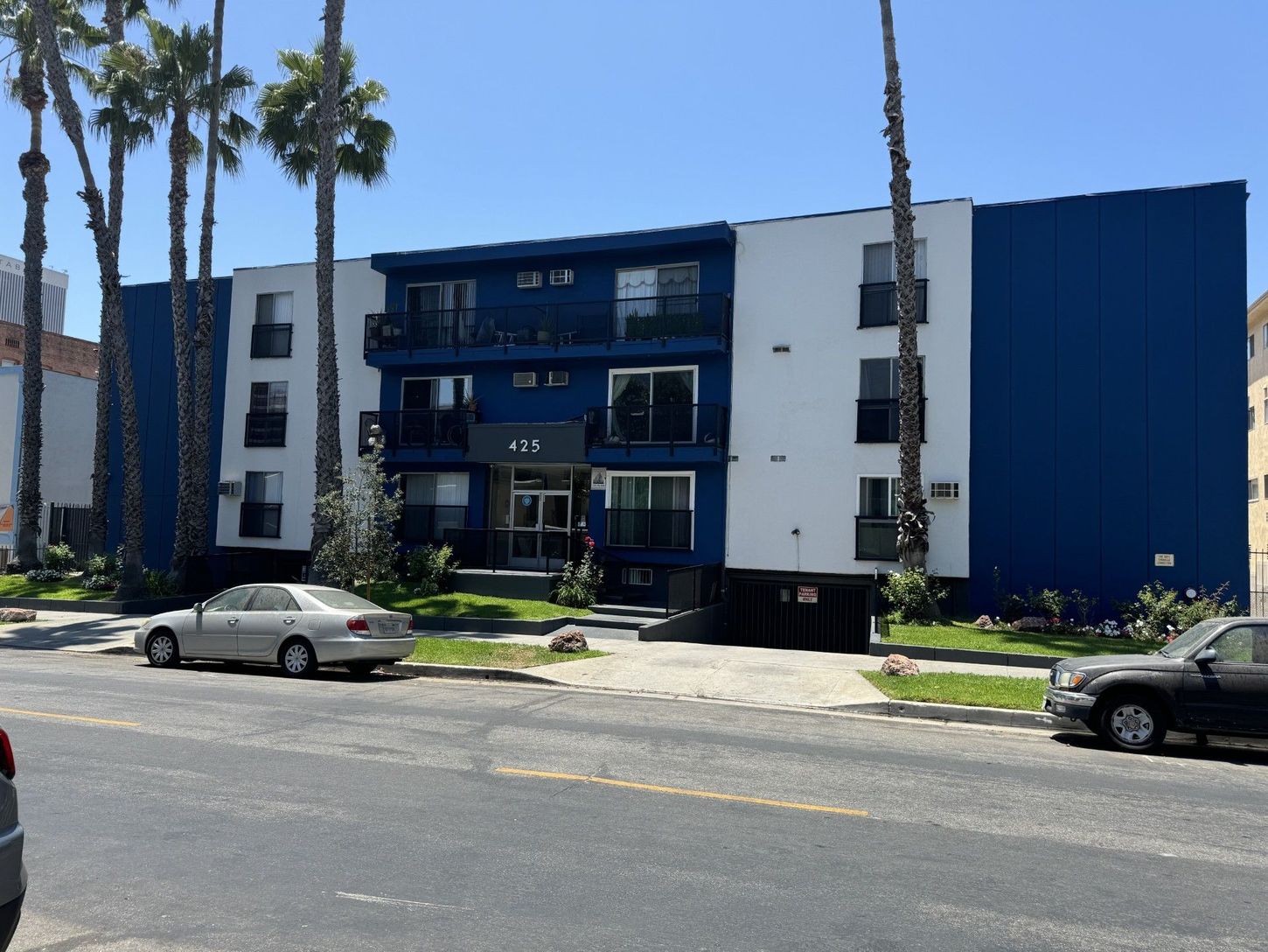 Apartment building with blue and white facade, palm trees, cars parked on street.