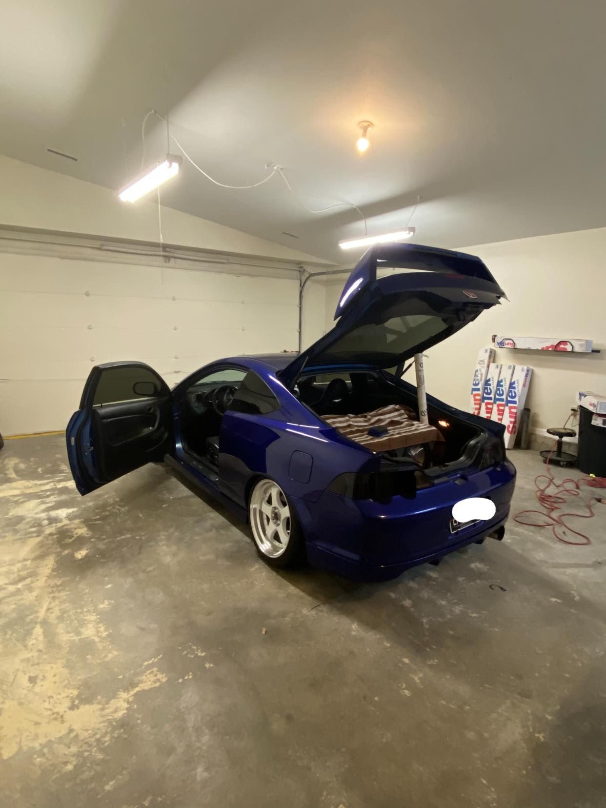 A dark blue sports car with doors and trunk open in a garage. It has white wheels and tinted windows.