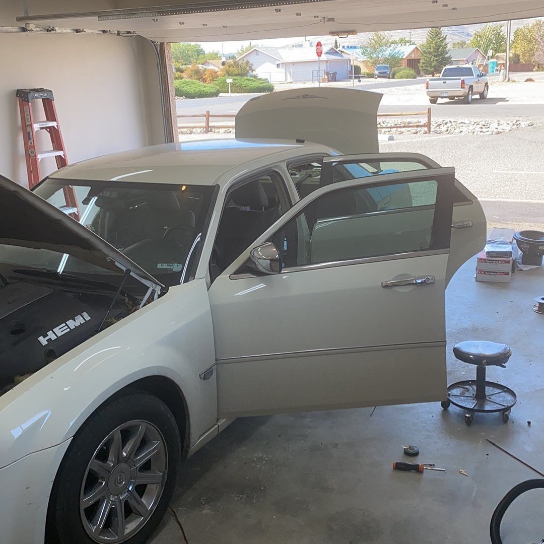 White car with doors open, parked under a carport, engine exposed; mechanic is possibly working on it.