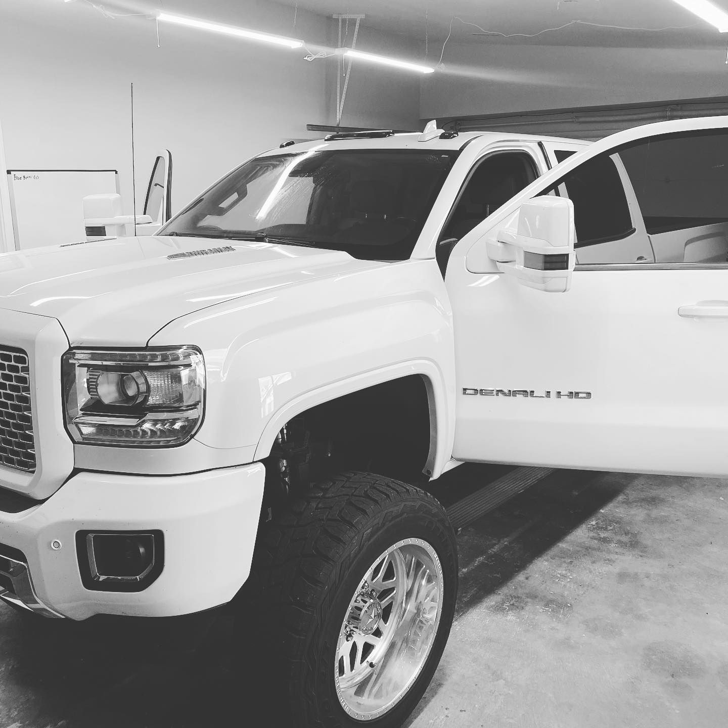 White lifted GMC Denali truck with open door, silver wheels, and tinted windows parked inside a garage.