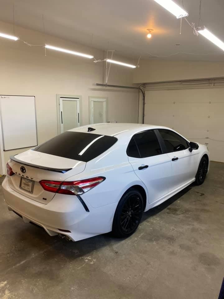 White Toyota Camry sedan with black wheels in a garage. Dark tinted windows and a rear spoiler.