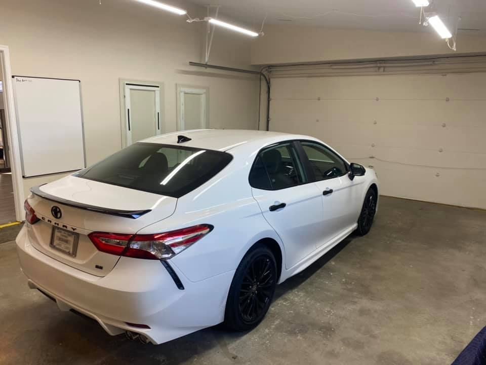 White Toyota Camry parked inside a garage. The car has black wheels and a black spoiler, with a license plate visible.