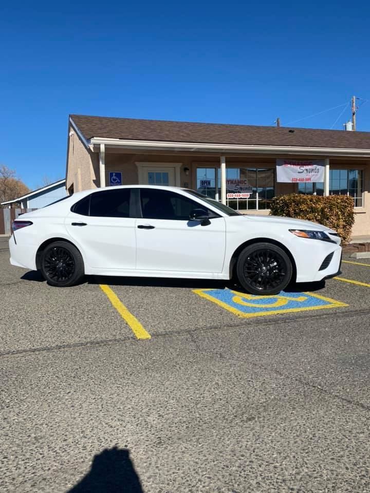 White Toyota Camry with black wheels parked in a handicap spot in front of a tan building on a sunny day.