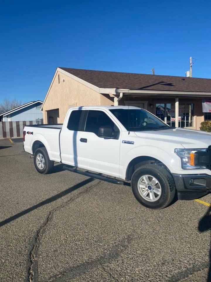 White Ford F-150 pickup truck parked on a cracked asphalt lot in front of a beige building. Blue sky overhead.