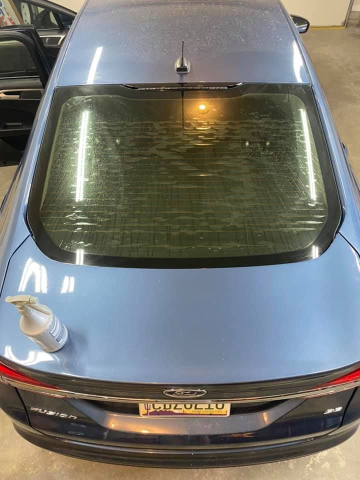 A light blue car with water-streaked rear window. A spray bottle sits on the car's trunk. The car is parked indoors.