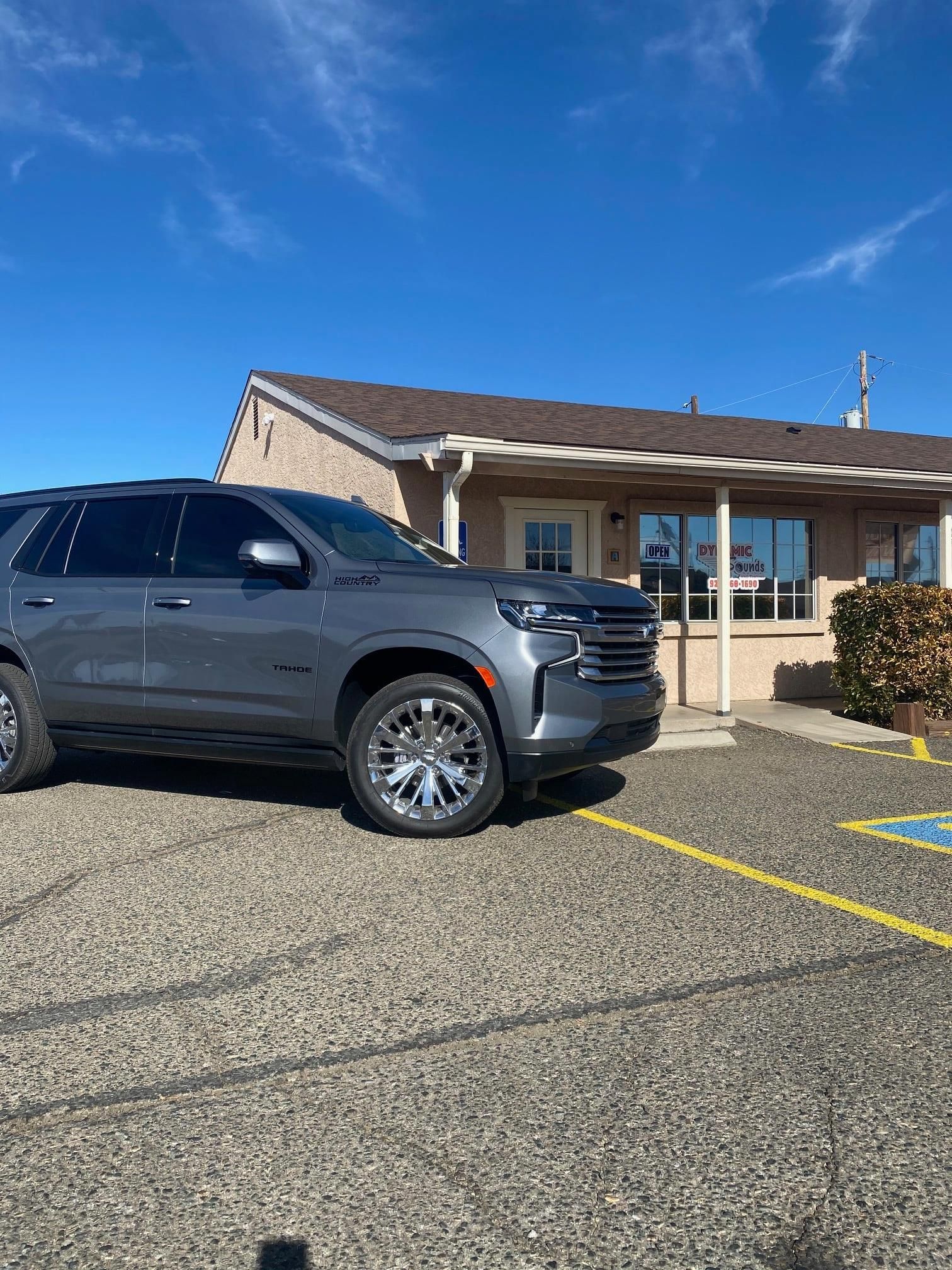 Gray SUV parked outside a beige building on a sunny day. The car has chrome wheels.