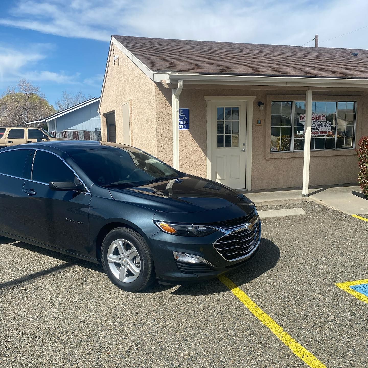Dark blue Chevrolet Malibu parked in front of a beige building with a white door and windows, under a bright blue sky.