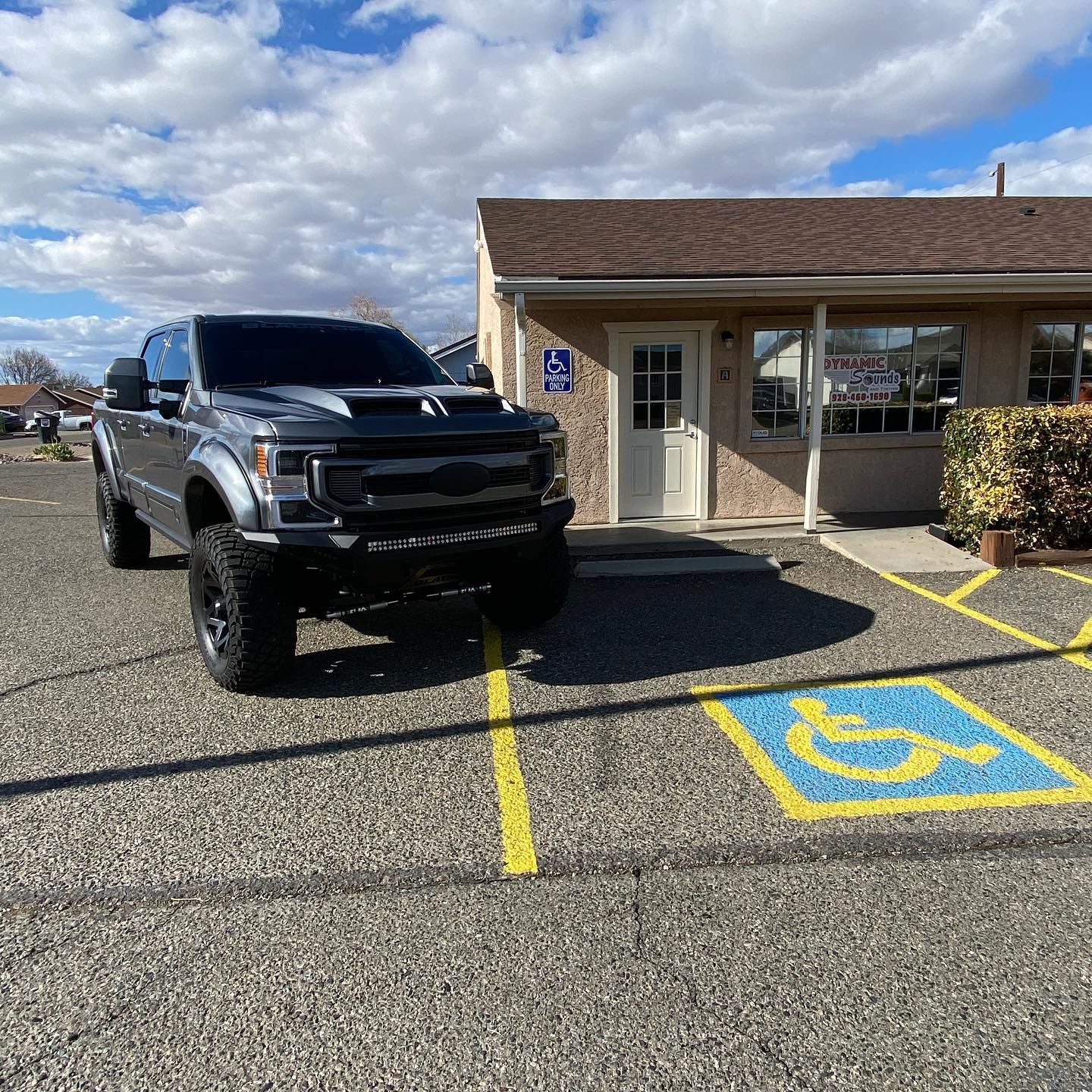 A large lifted gray truck parked in a handicapped parking spot in front of a small business building. Cloudy day.