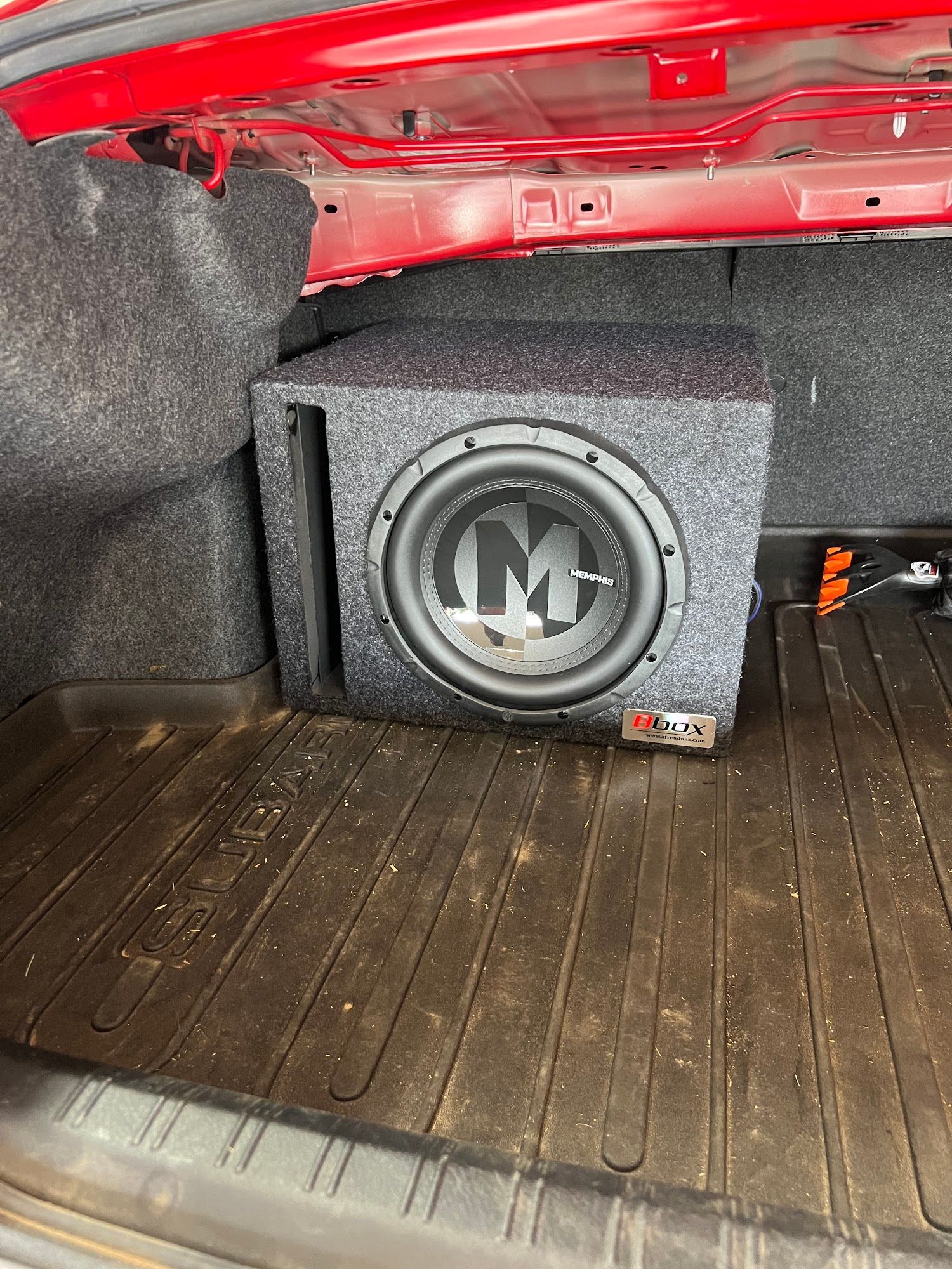 A subwoofer box with a large speaker is in the trunk of a red car, sitting on a rubber mat.