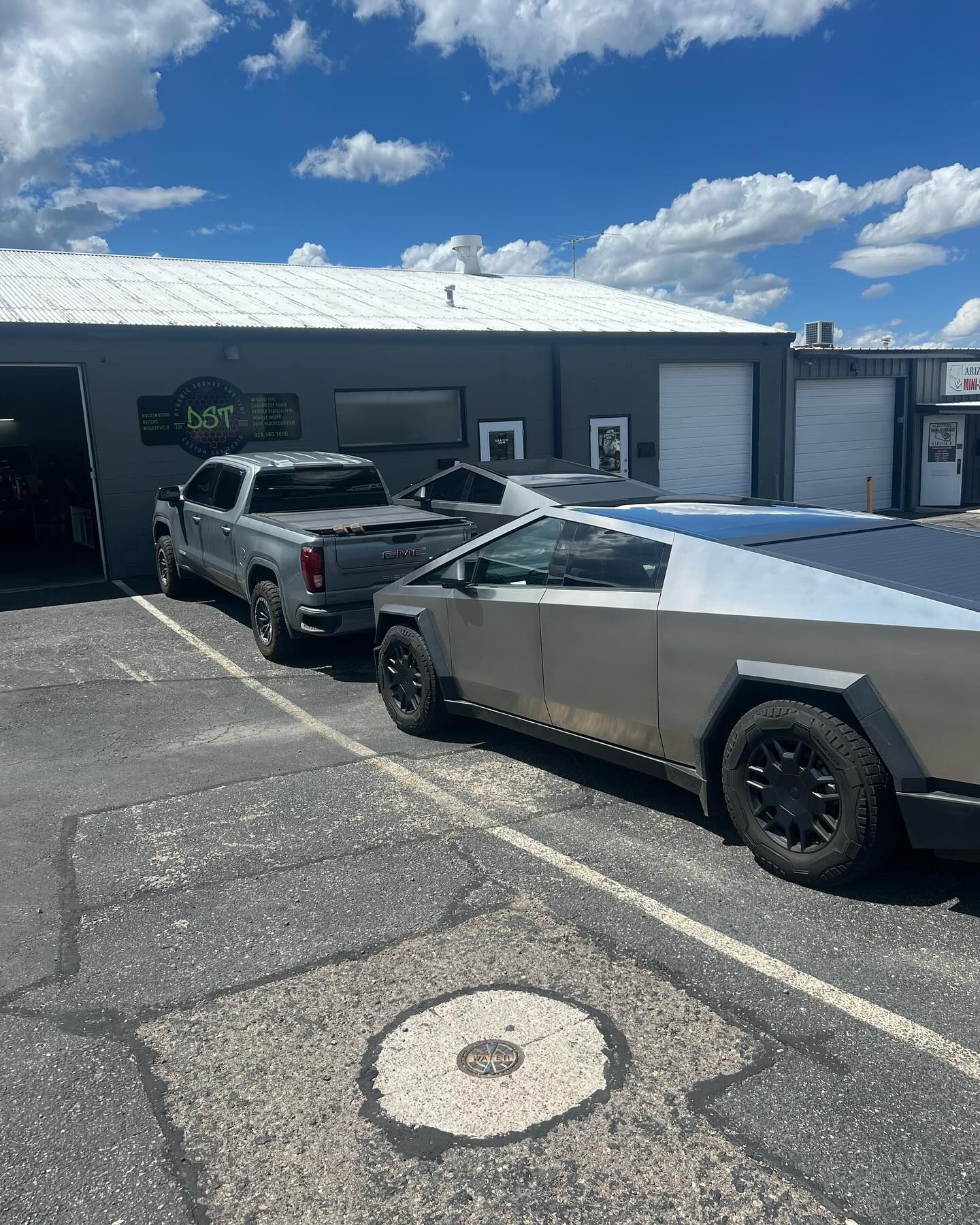 A silver Tesla Cybertruck and a gray pickup truck parked outside a building under a blue sky.