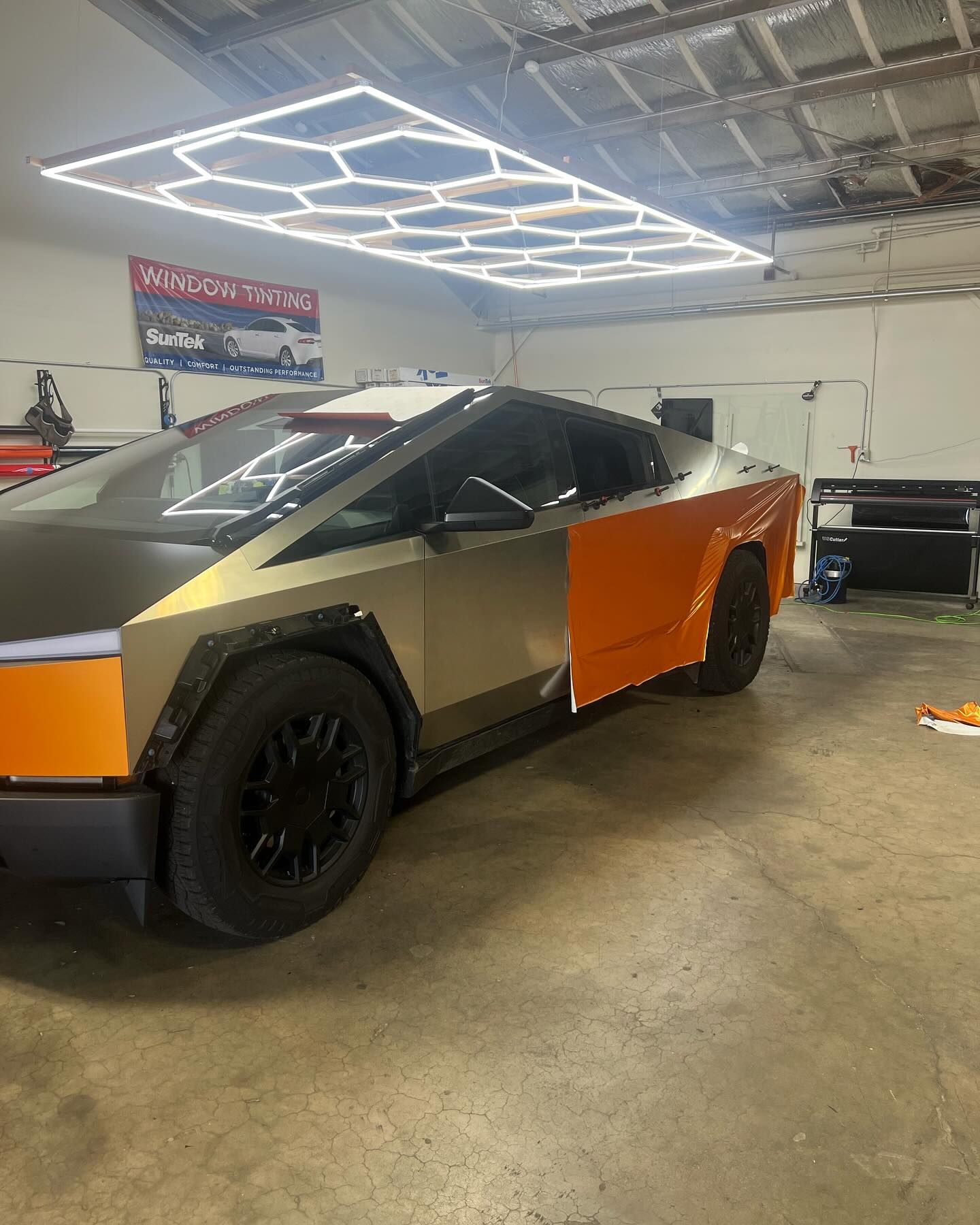 Tesla Cybertruck with a partial orange and gold wrap in a garage with hexagon lights.