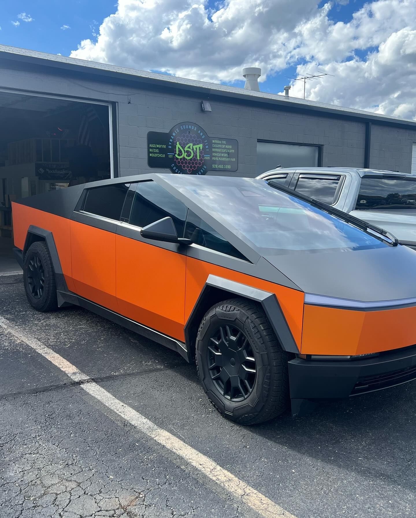 Tesla Cybertruck, orange and gray wrap, parked in front of a business with a partially open garage door on a sunny day.