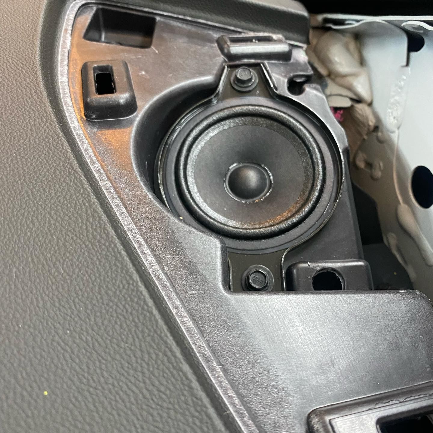 A car's dashboard with a small, round speaker installed. The speaker is black, positioned in a molded plastic housing.