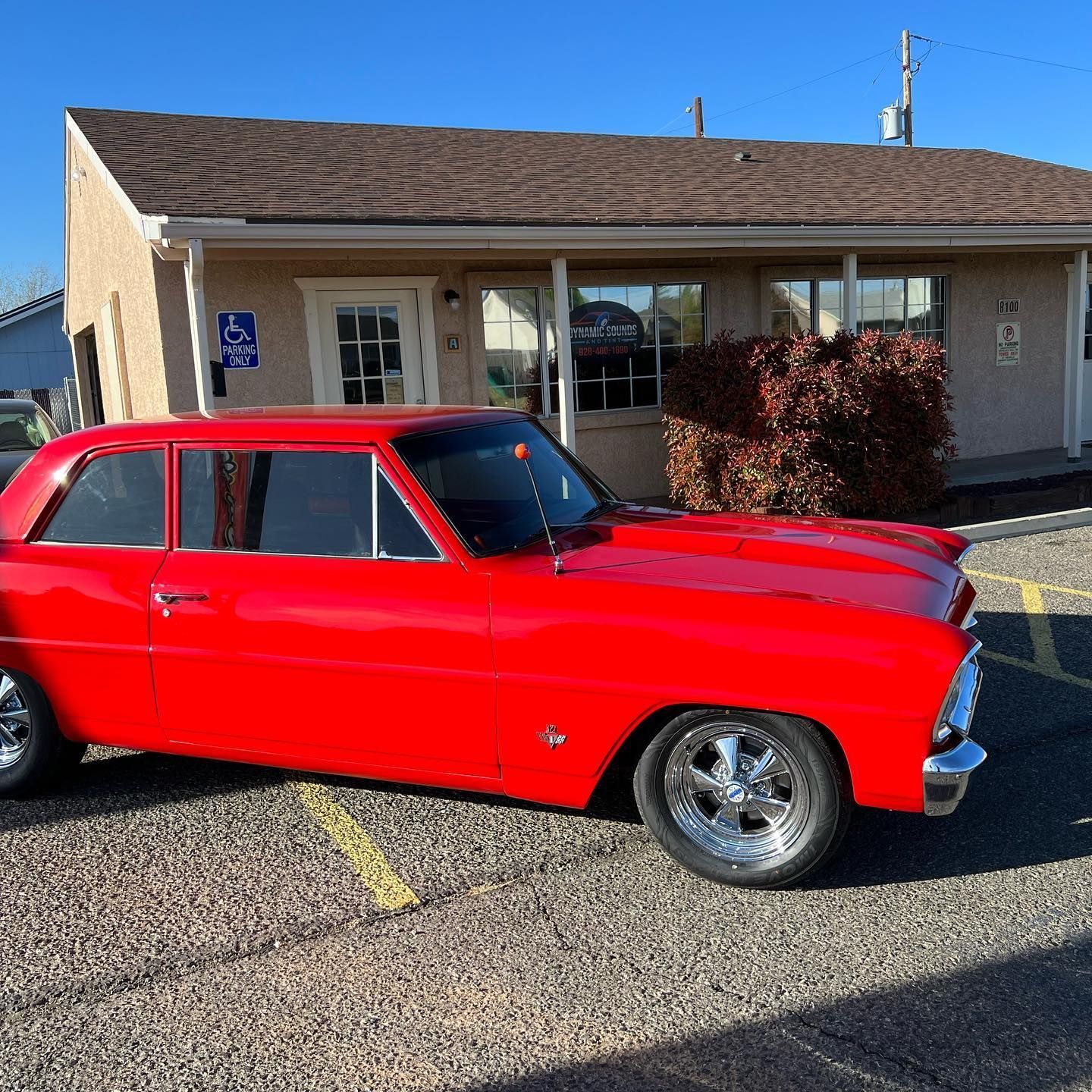 Bright red classic car parked in front of a tan-colored building with a brown roof, on a sunny day.