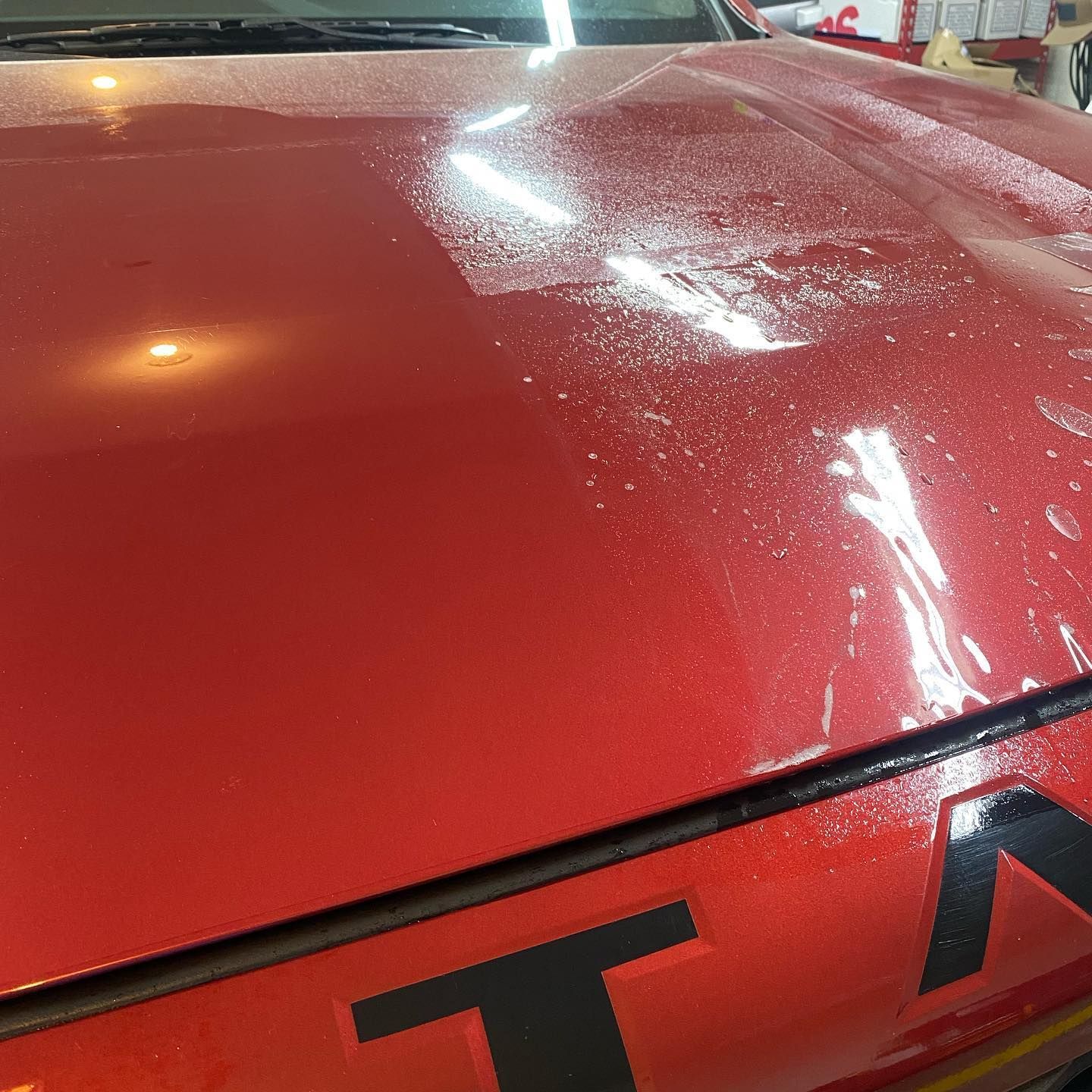 Red car hood with water droplets and part of a black logo; likely after a car wash or application.