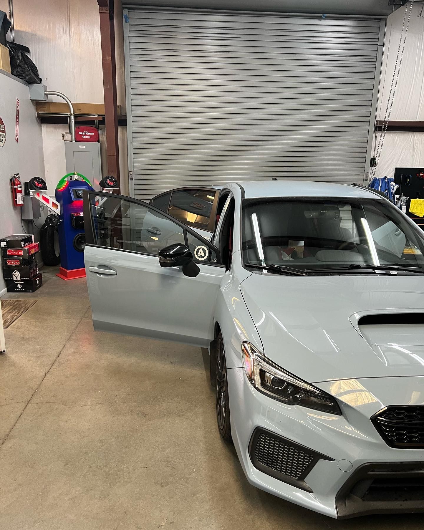 A light blue Subaru WRX with the driver's side door open, inside a garage. The garage door is closed.