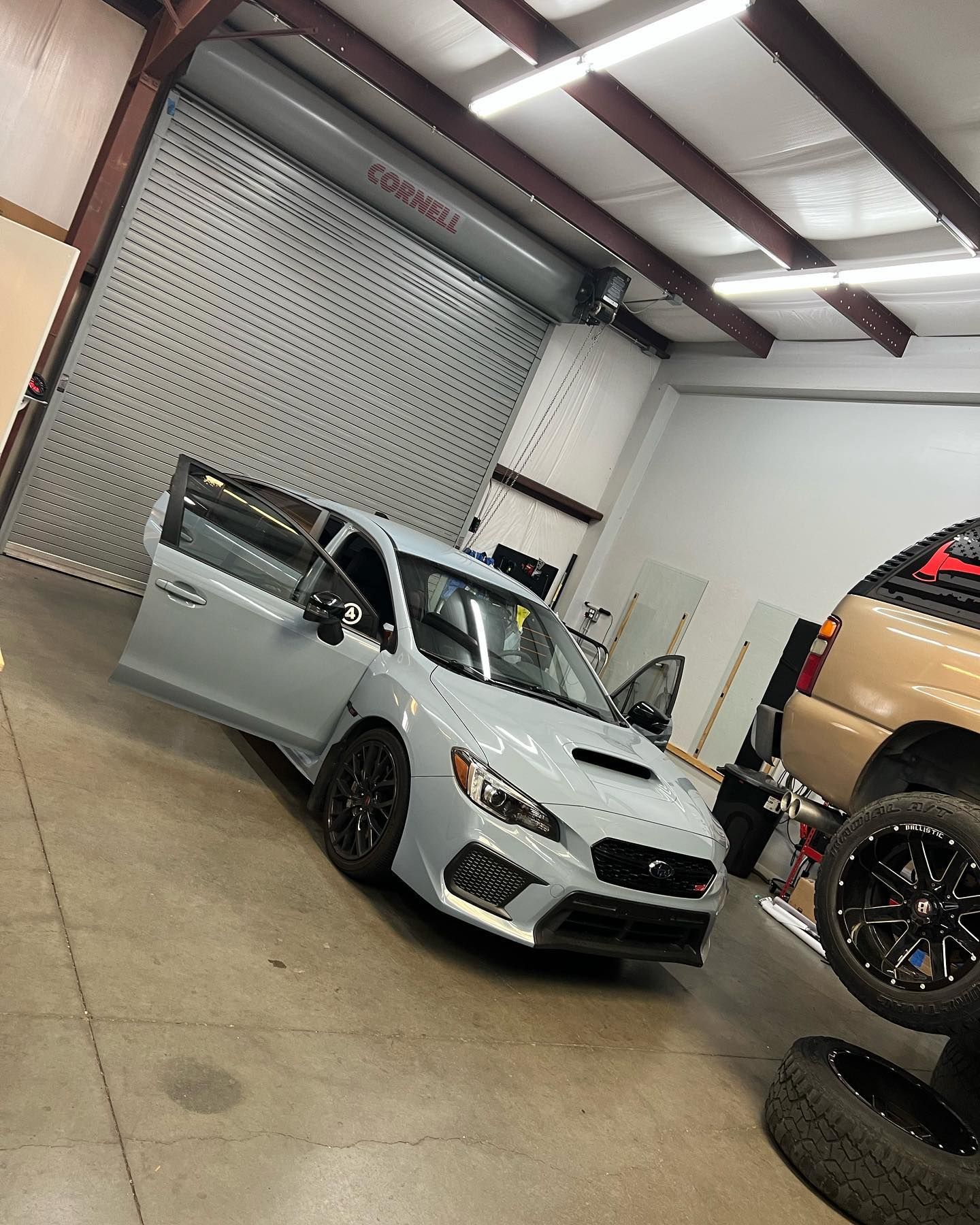 Light gray Subaru WRX with open doors inside a garage with a closed gray garage door and a tan SUV in the background.