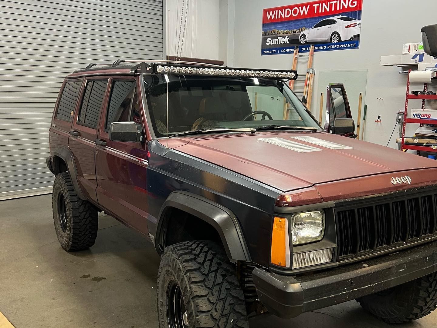 A modified maroon Jeep Cherokee with black wheels and a light bar in a garage, likely for window tinting.