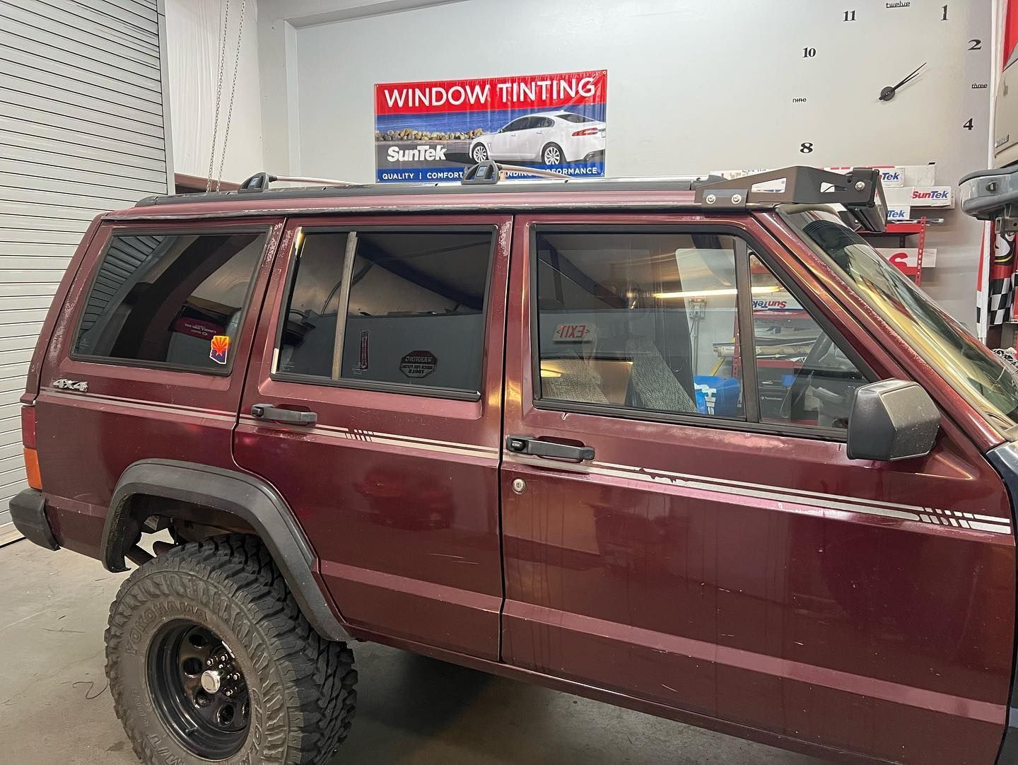 A maroon Jeep Cherokee with tinted windows parked inside a window tinting shop. Roof rack and off-road tires are visible.