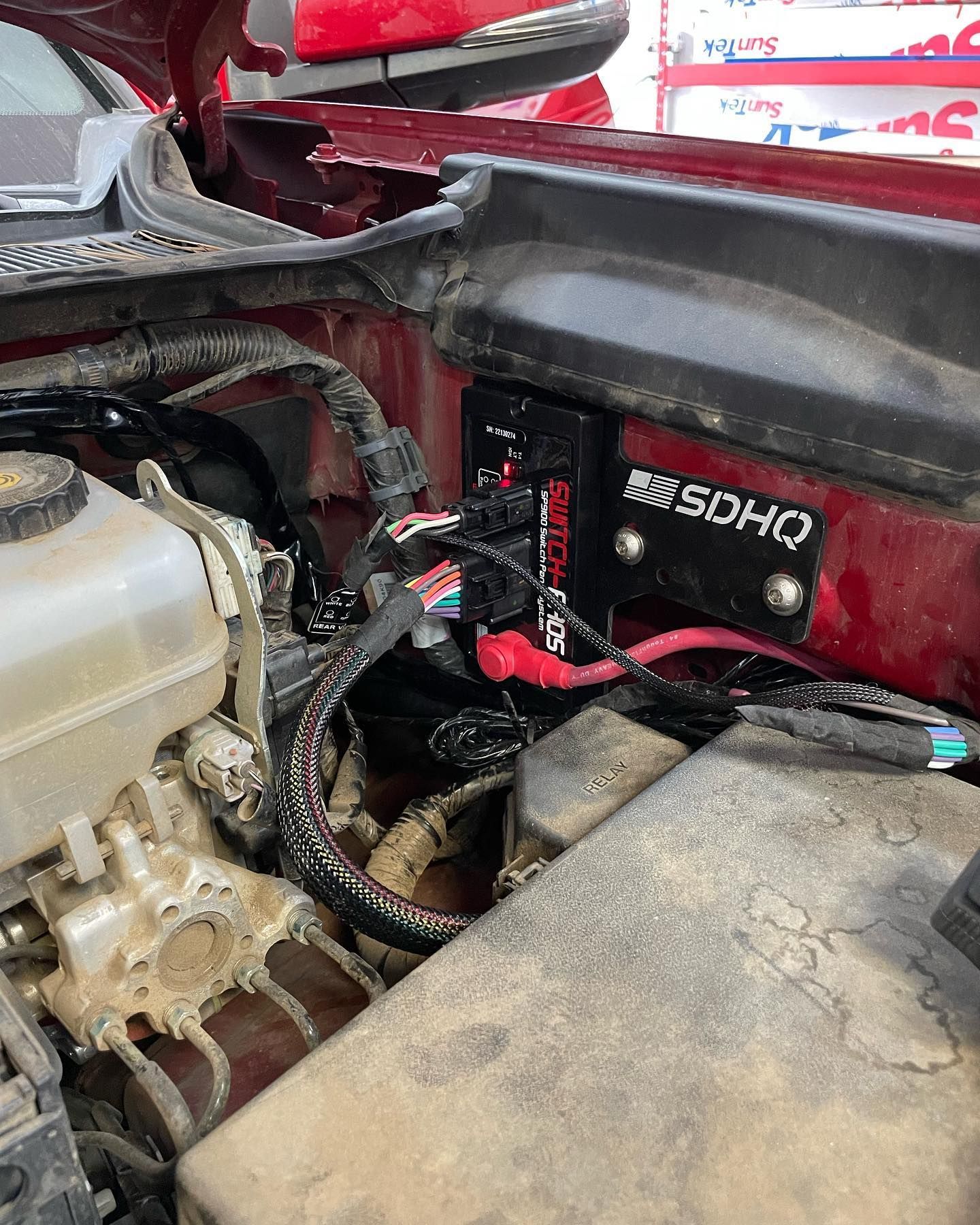 Black SDHQ power distribution box mounted in a red vehicle's engine bay, surrounded by wires and components.