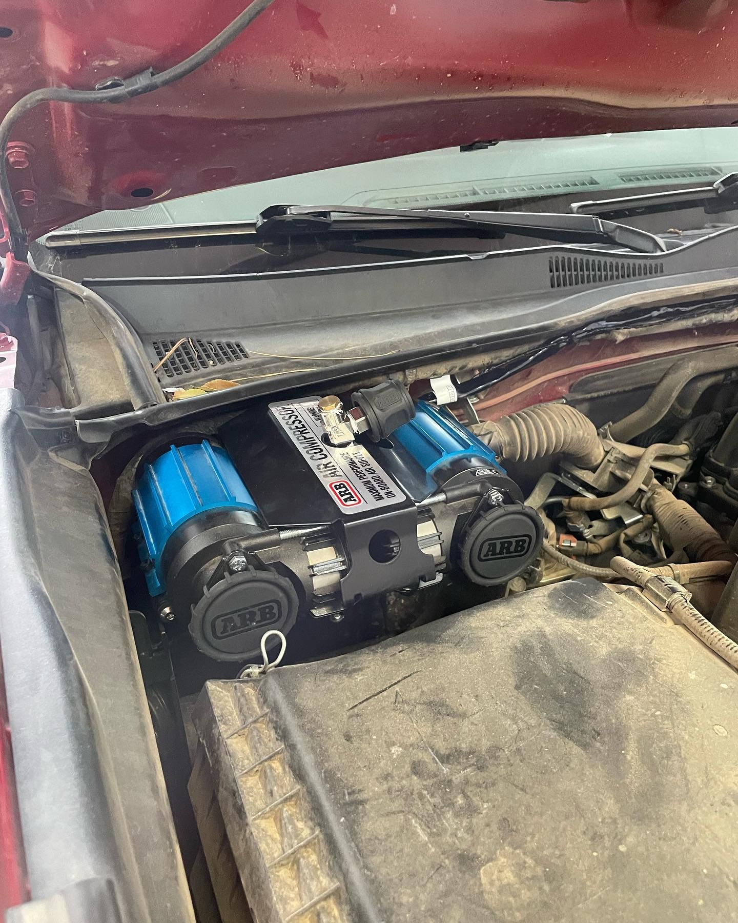 Blue air compressor mounted in a vehicle's engine bay, next to the windshield. The compressor is black with blue air filters.