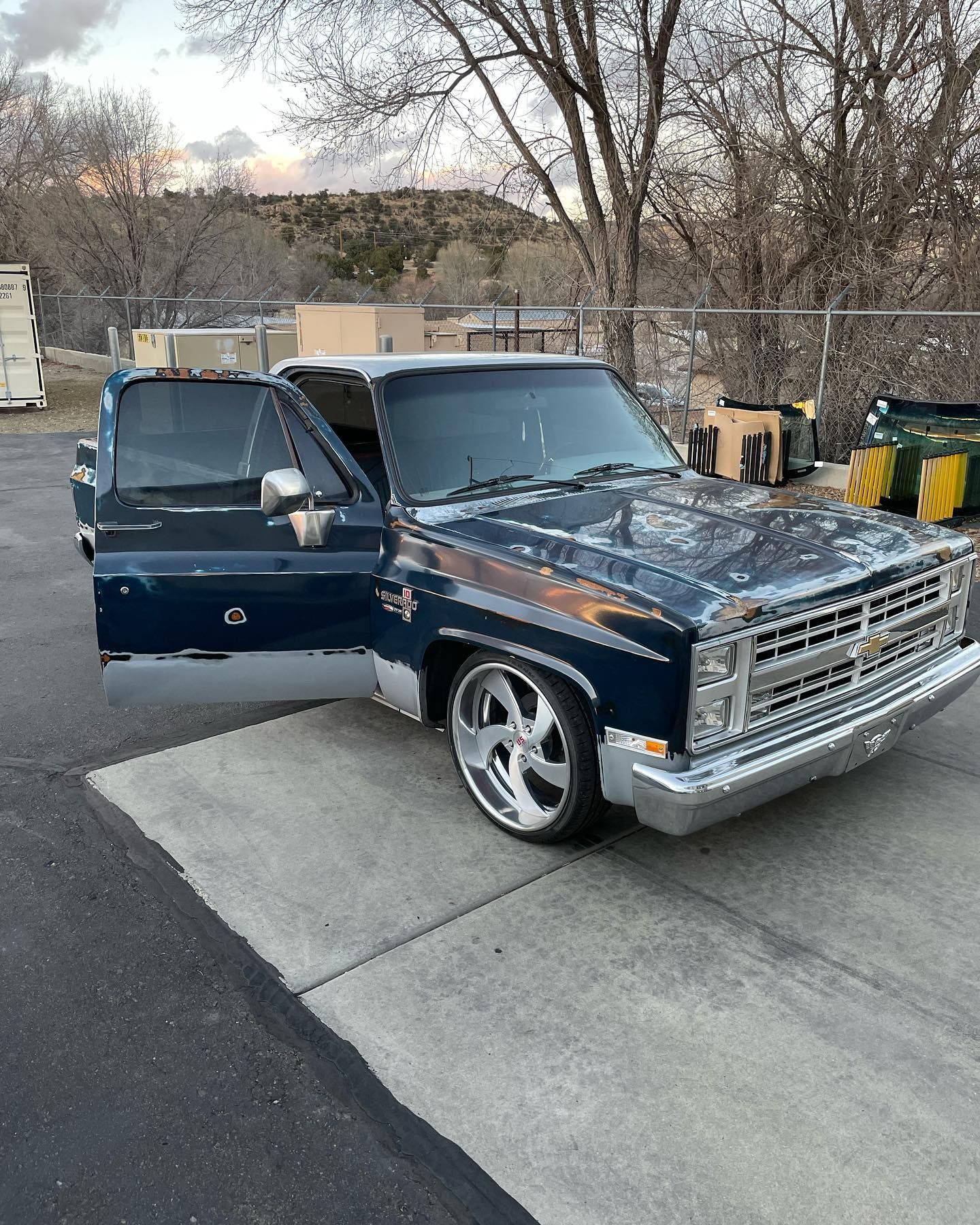 Blue Chevrolet pickup truck with open door, lowered suspension, chrome wheels, parked on concrete near trees.