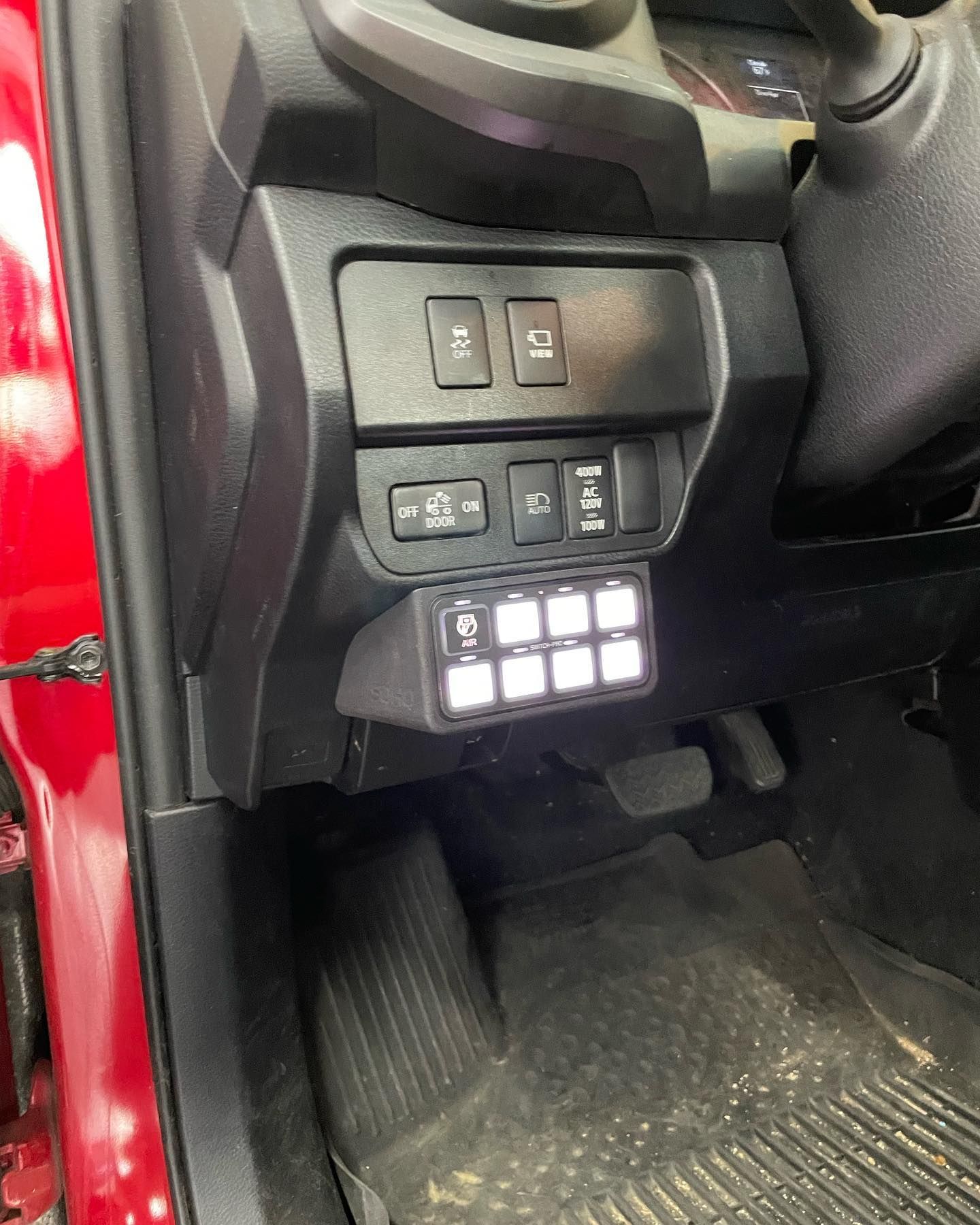 Interior shot of a red car's dashboard. A switch panel with illuminated white buttons is mounted below existing switches.