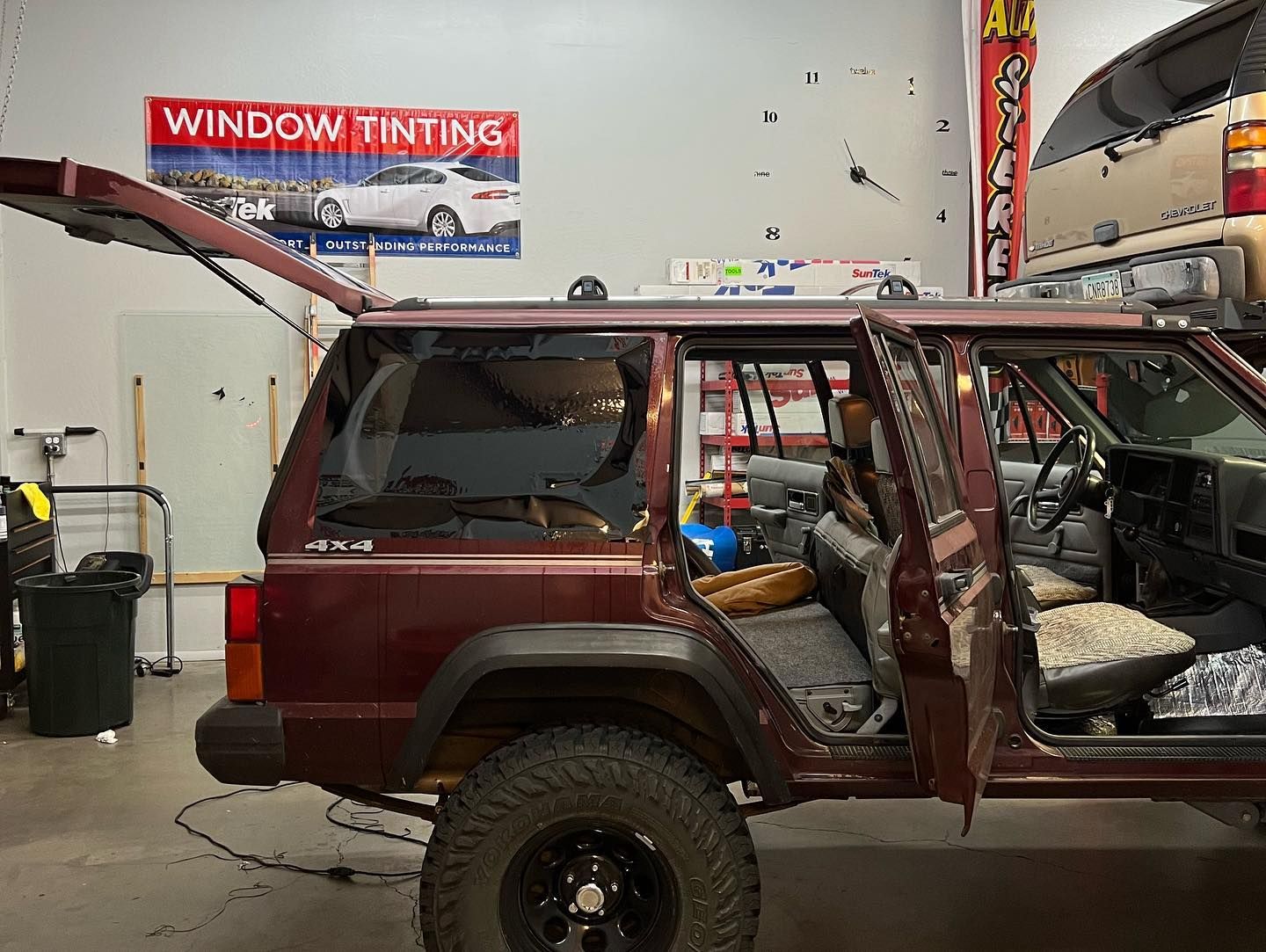 A maroon Jeep Cherokee with its doors open, parked in a garage; window tinting sign is on the wall.