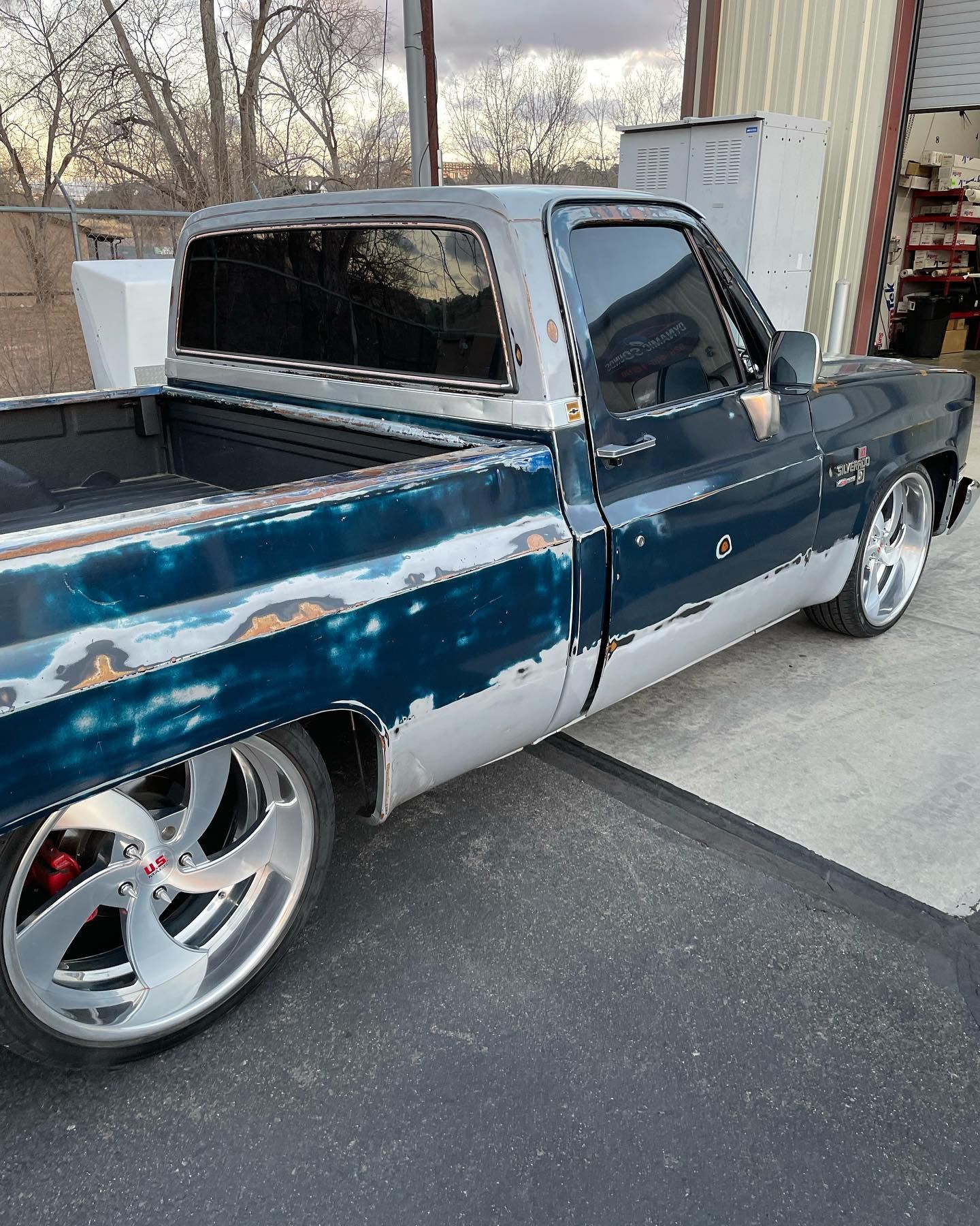 Classic blue and silver pickup truck with large custom wheels, parked outside a garage. The paint shows signs of wear.