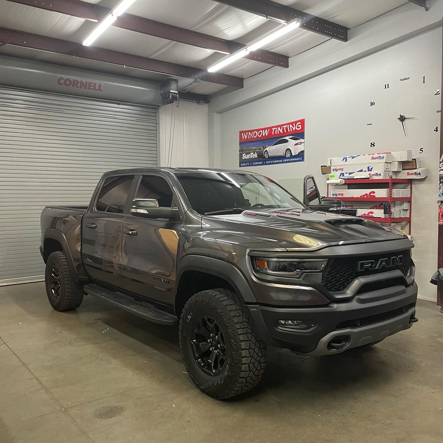 A gray Ram TRX truck with camouflage accents is inside a garage, near a roll-up door and shelves of materials.