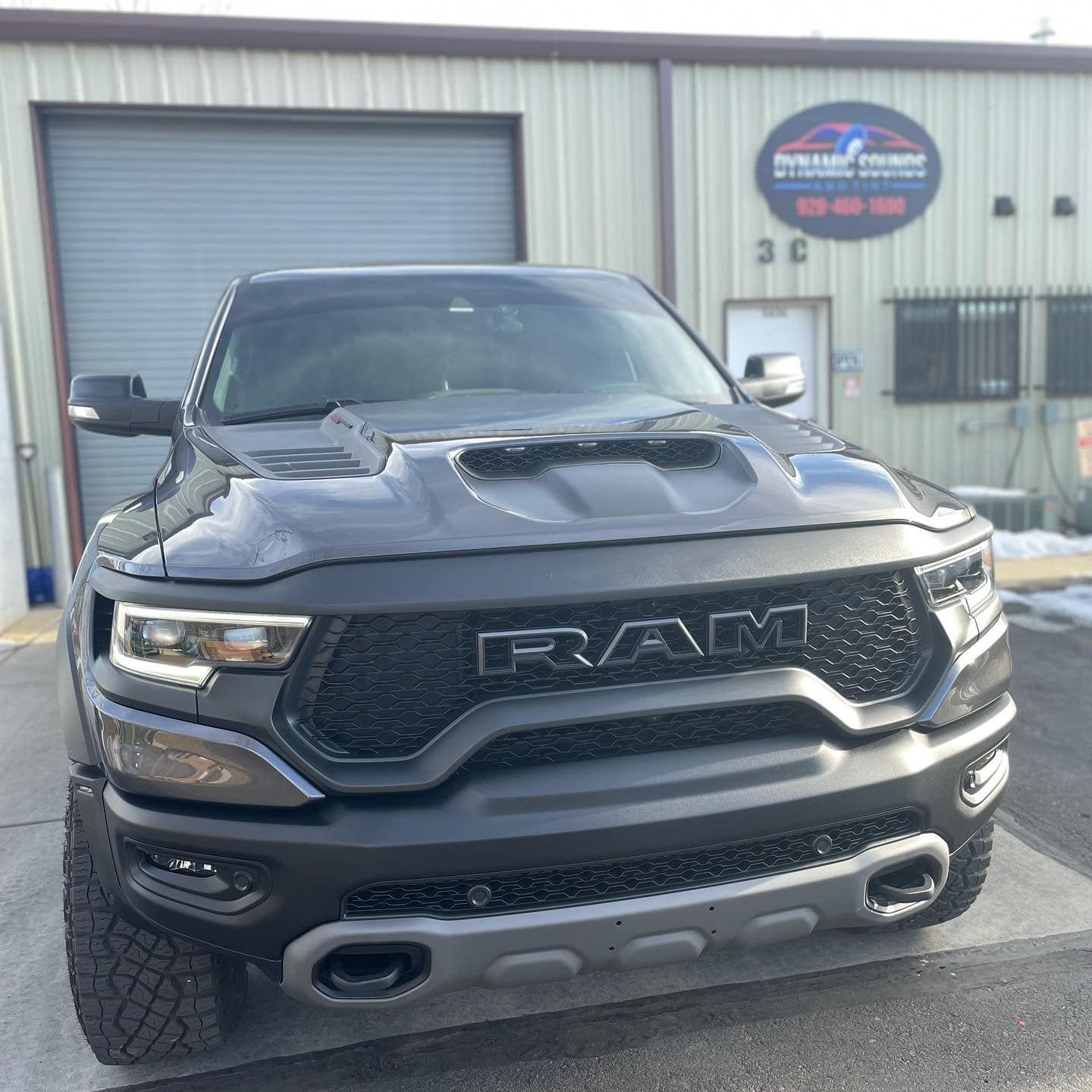 A gray Ram TRX pickup truck parked in front of a garage with a sign reading 