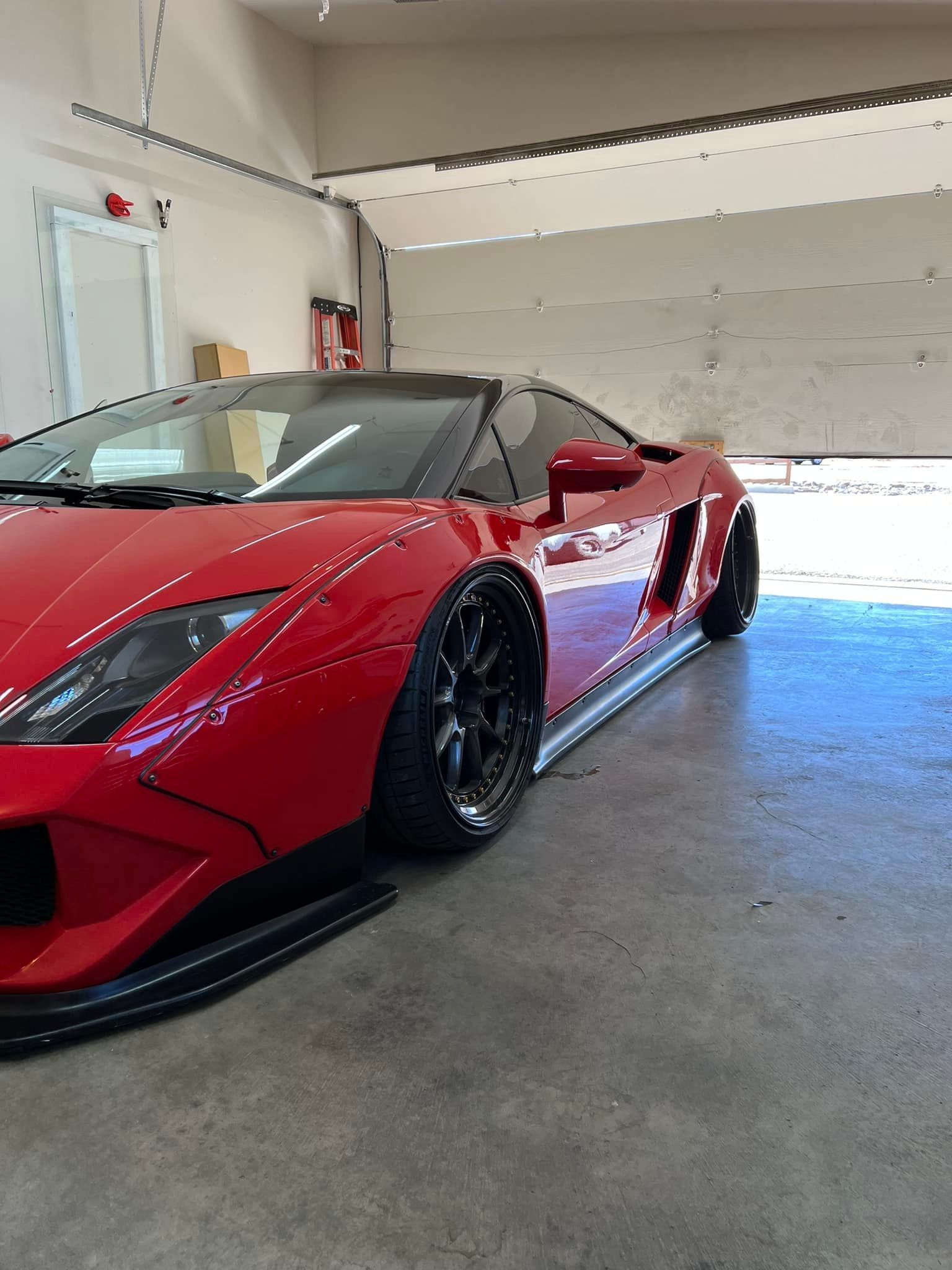 Red Lamborghini sports car parked in a garage with the door open. Black rims, side skirts, and a black roof.