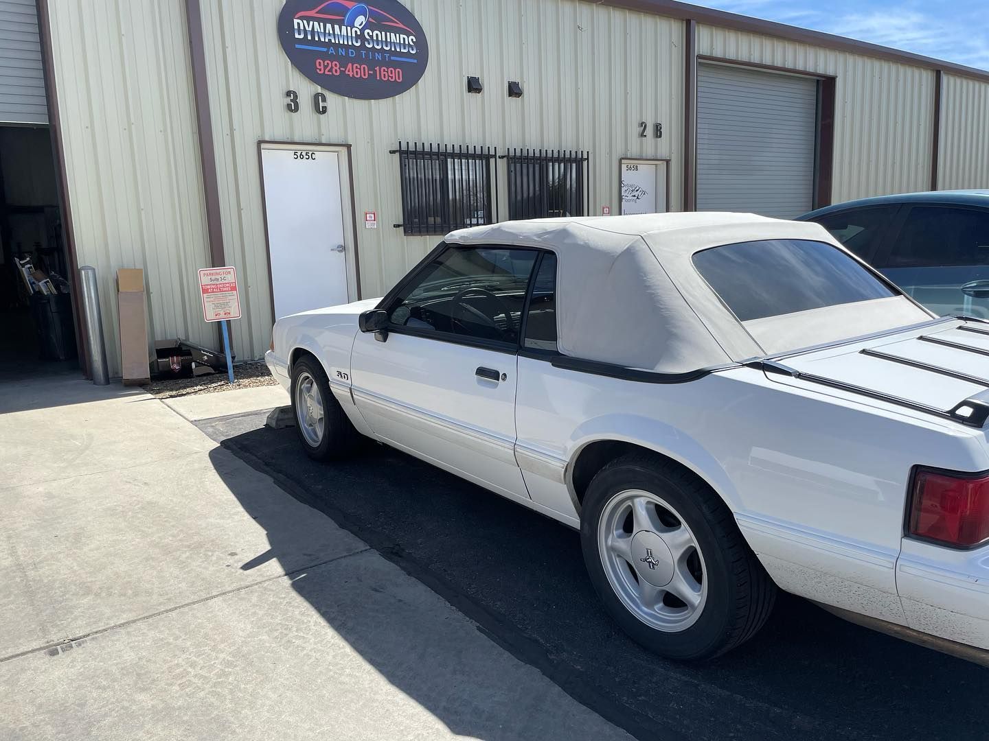 White Ford Mustang convertible parked outside a light-colored building with a sign. The car's top is up, and the day is sunny.