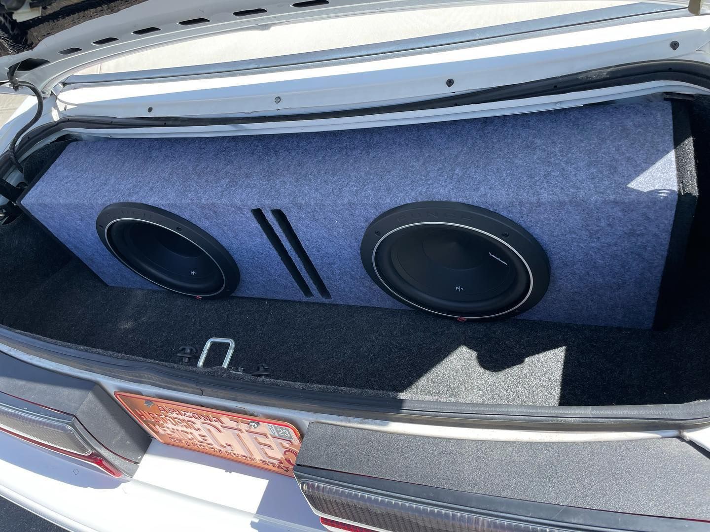 A car trunk with a gray, dual-subwoofer speaker box installed. The box is covered in gray fabric.