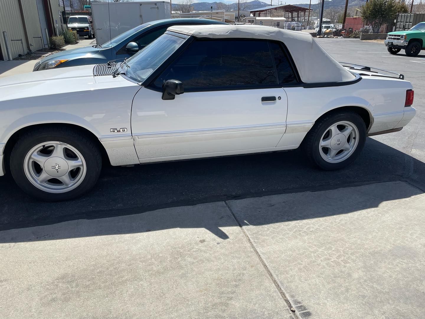 White Ford Mustang convertible parked on a gray surface with a white convertible top, surrounded by other vehicles.