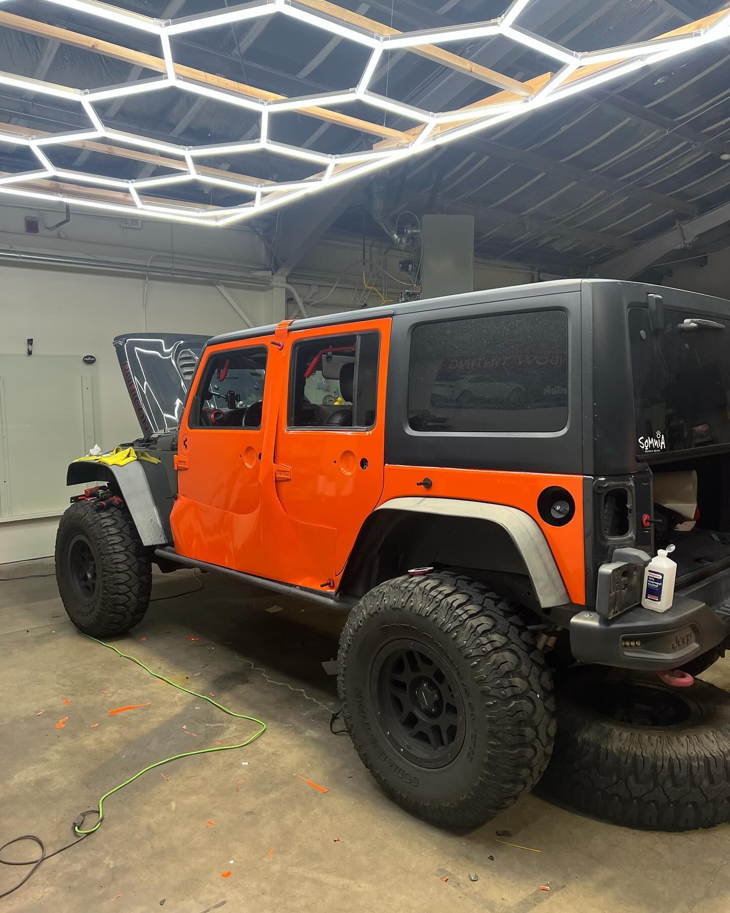 A partially orange Jeep Wrangler sits inside a garage under hexagonal lights. The hood is up, with the tire removed on the right.