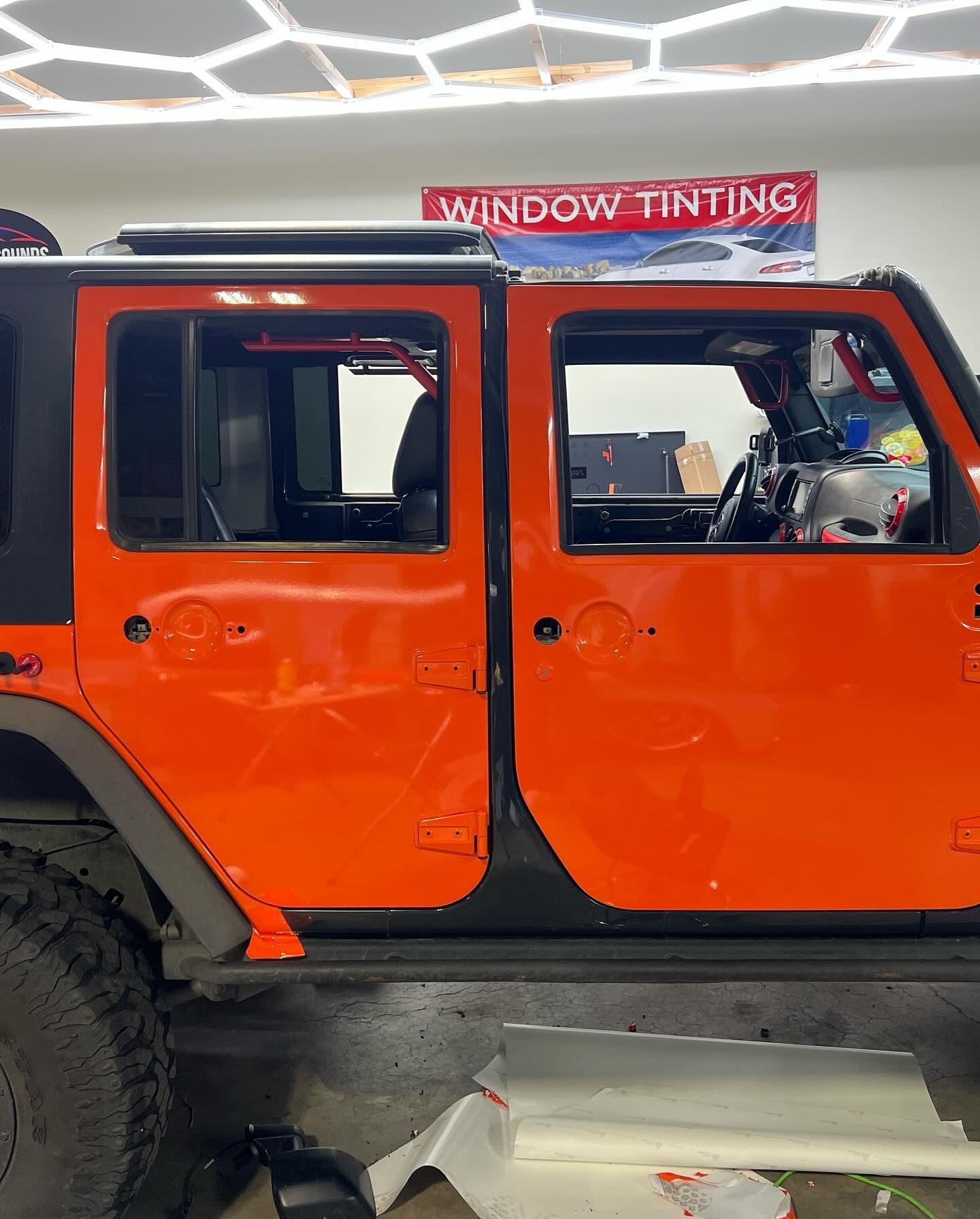 An orange Jeep Wrangler with tinted windows is in a window tinting shop. A 