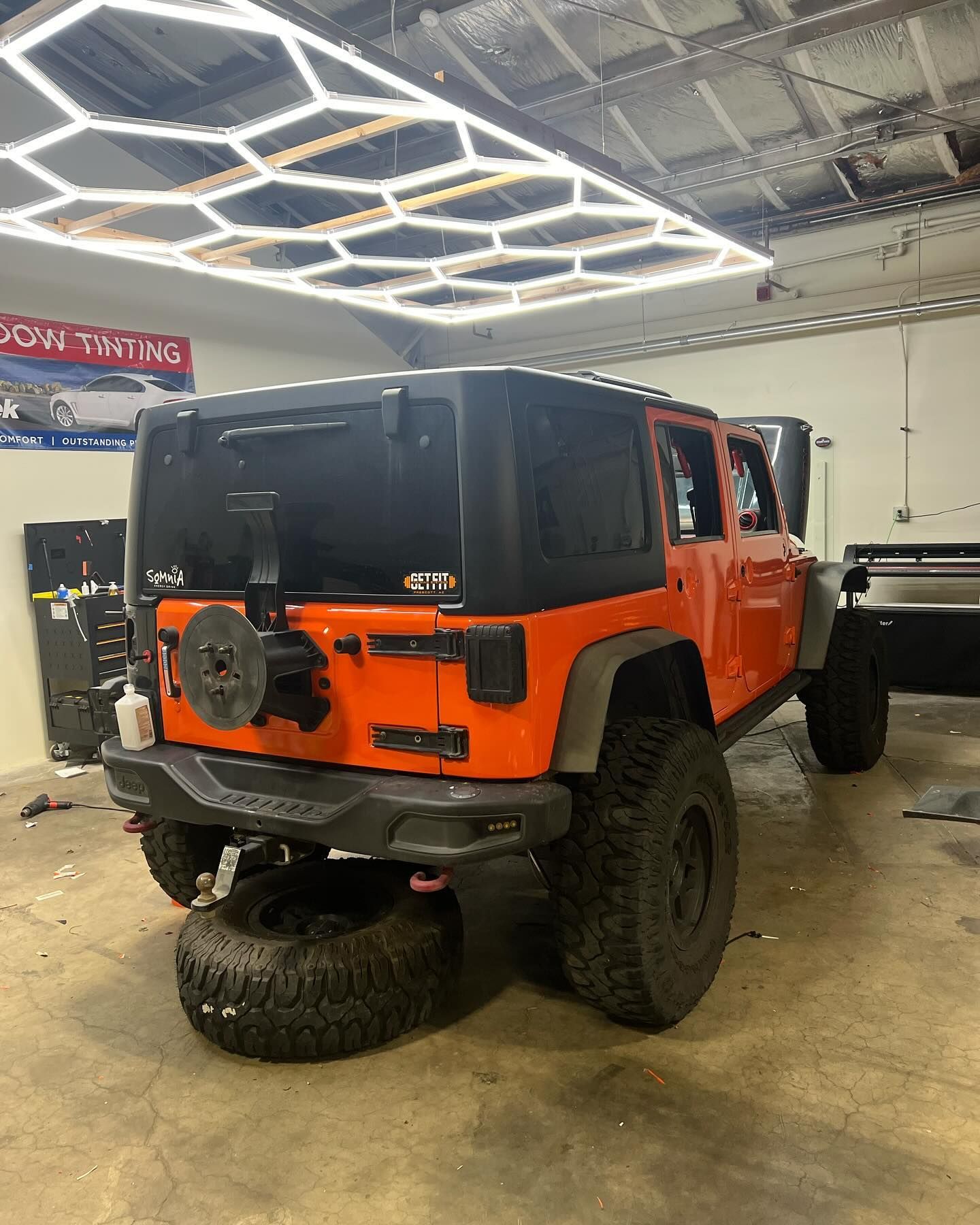 Orange Jeep Wrangler with black top in a shop, spare tire on the ground, under hexagonal lights.