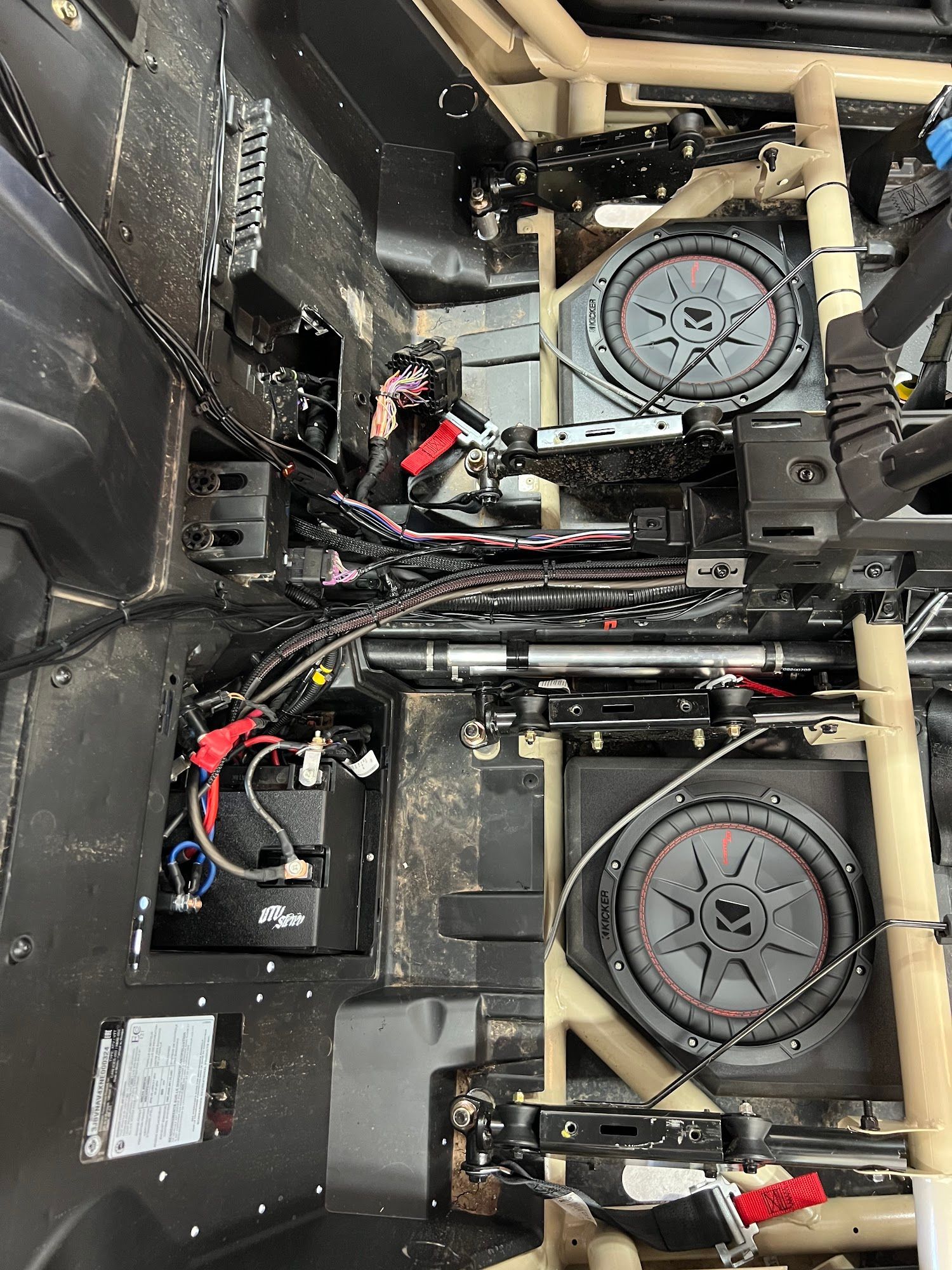 Interior of a vehicle with two large subwoofers installed. Wiring, black paneling, and a metal frame are visible.