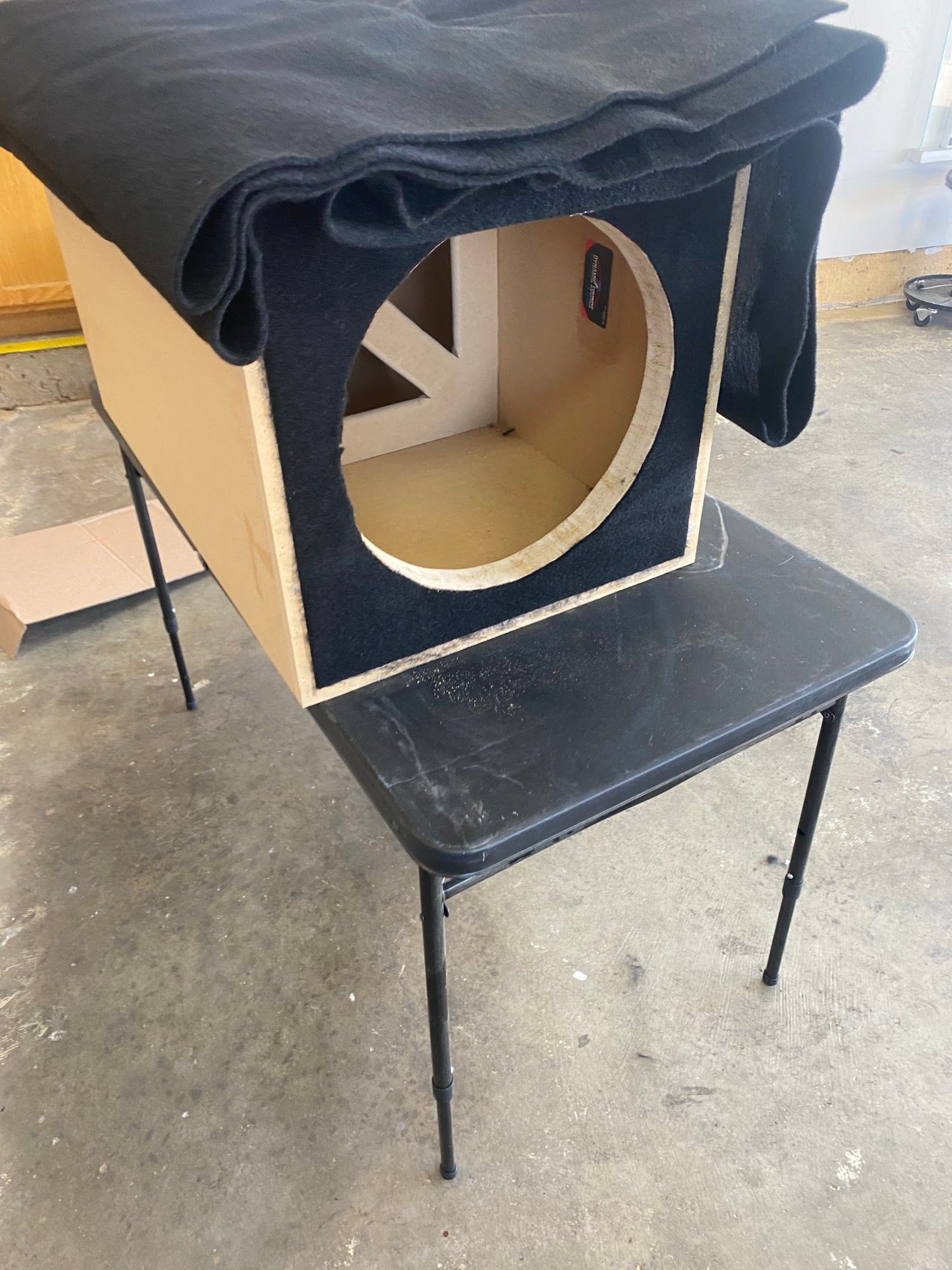 A subwoofer box made of light wood, with a circular cutout, sits on a black folding table. Black carpet covers the top.