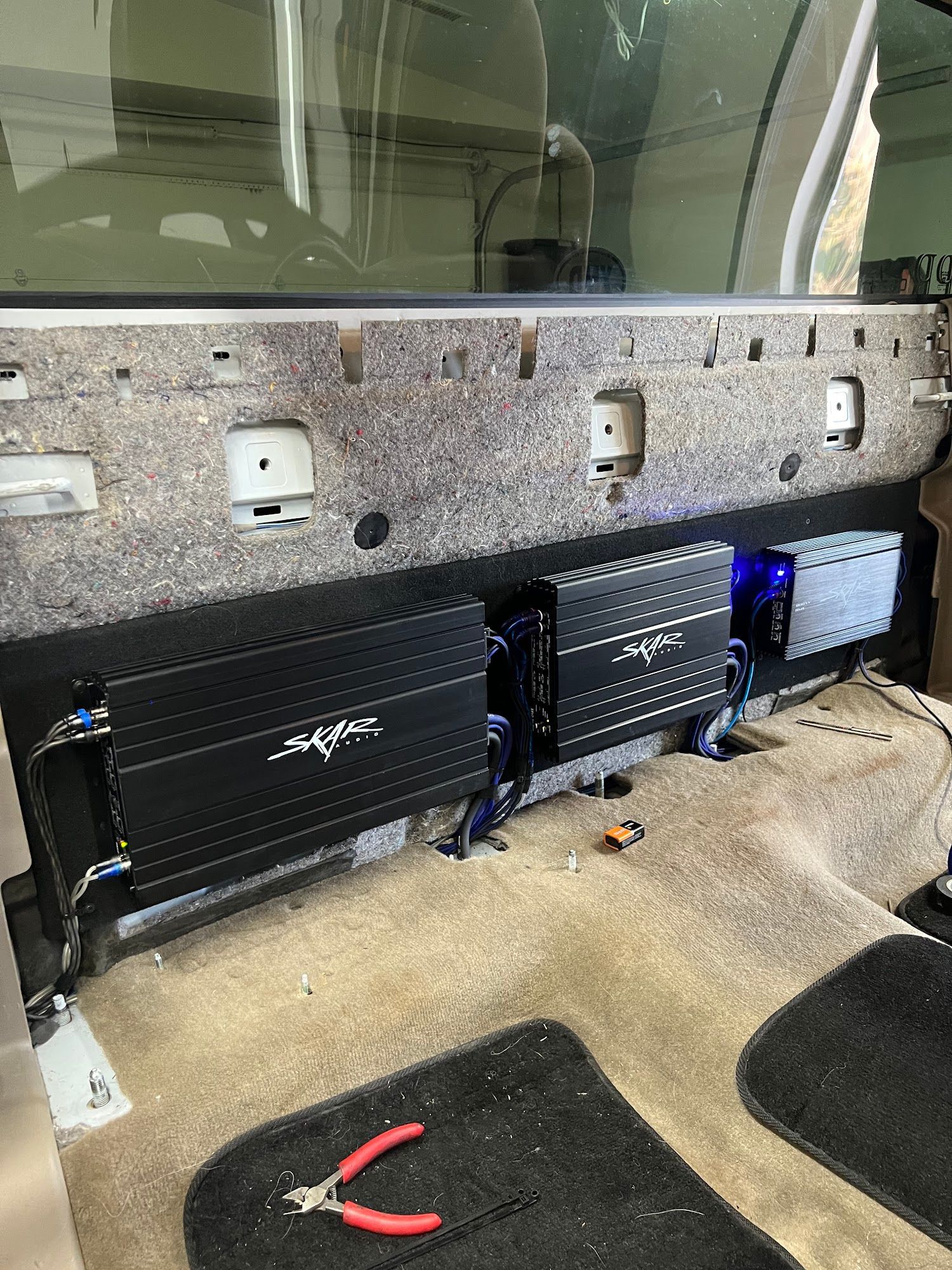 Car audio amplifier installation. Three black amplifiers are mounted on a back panel inside a vehicle. Carpet and tools are visible.